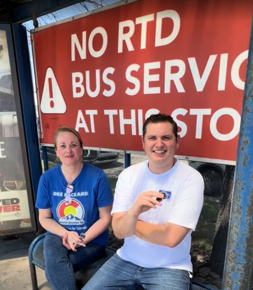 Two people sitting at a bus stop in front of a red sign that reads 'No RTD Bus Service at This Stop'.