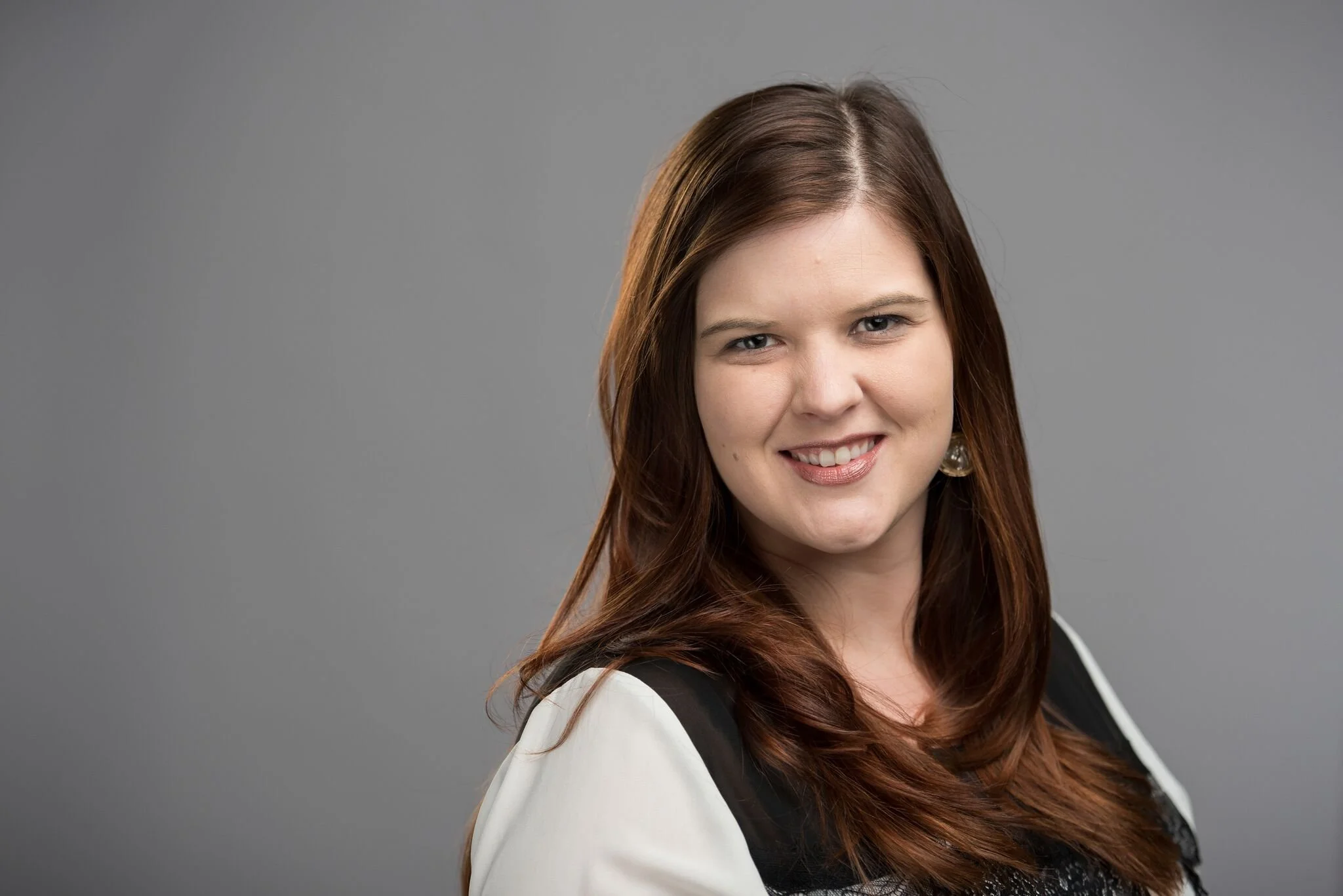 A portrait of Gena Ozols young woman with long brown hair, smiling, wearing a black and white top and earrings, against a plain gray background.