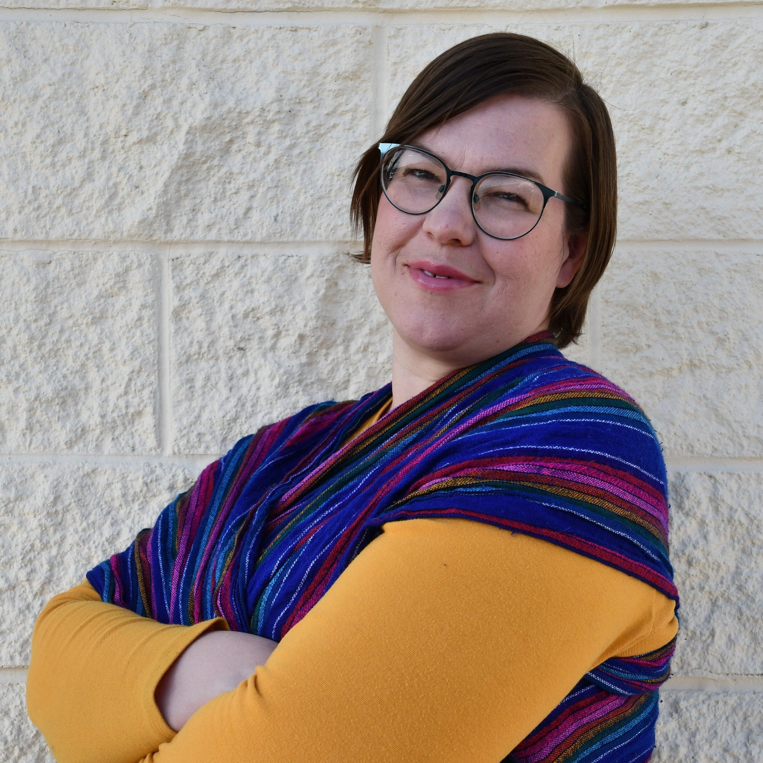 Alison Coombs, a woman with short brown hair, glasses, and light skin standing against a beige brick wall, smiling with her arms crossed, wearing a mustard yellow long sleeve shirt and a colorful striped shawl.