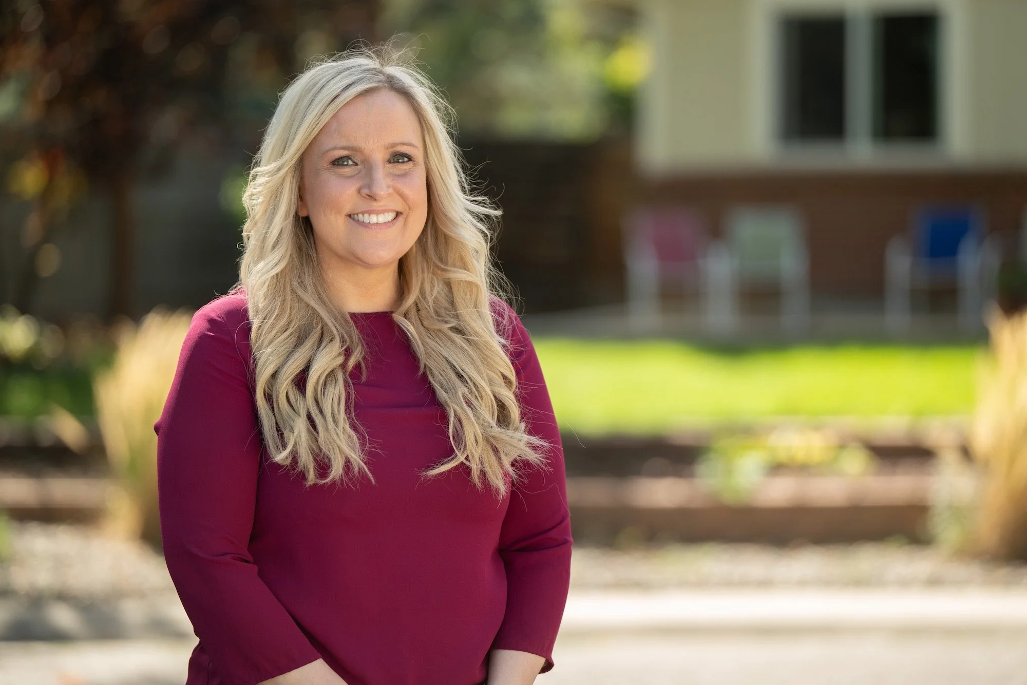 Gretchen Rydin smiling blonde woman in a maroon long-sleeve top standing outdoors in front of a house with green lawn and trees.