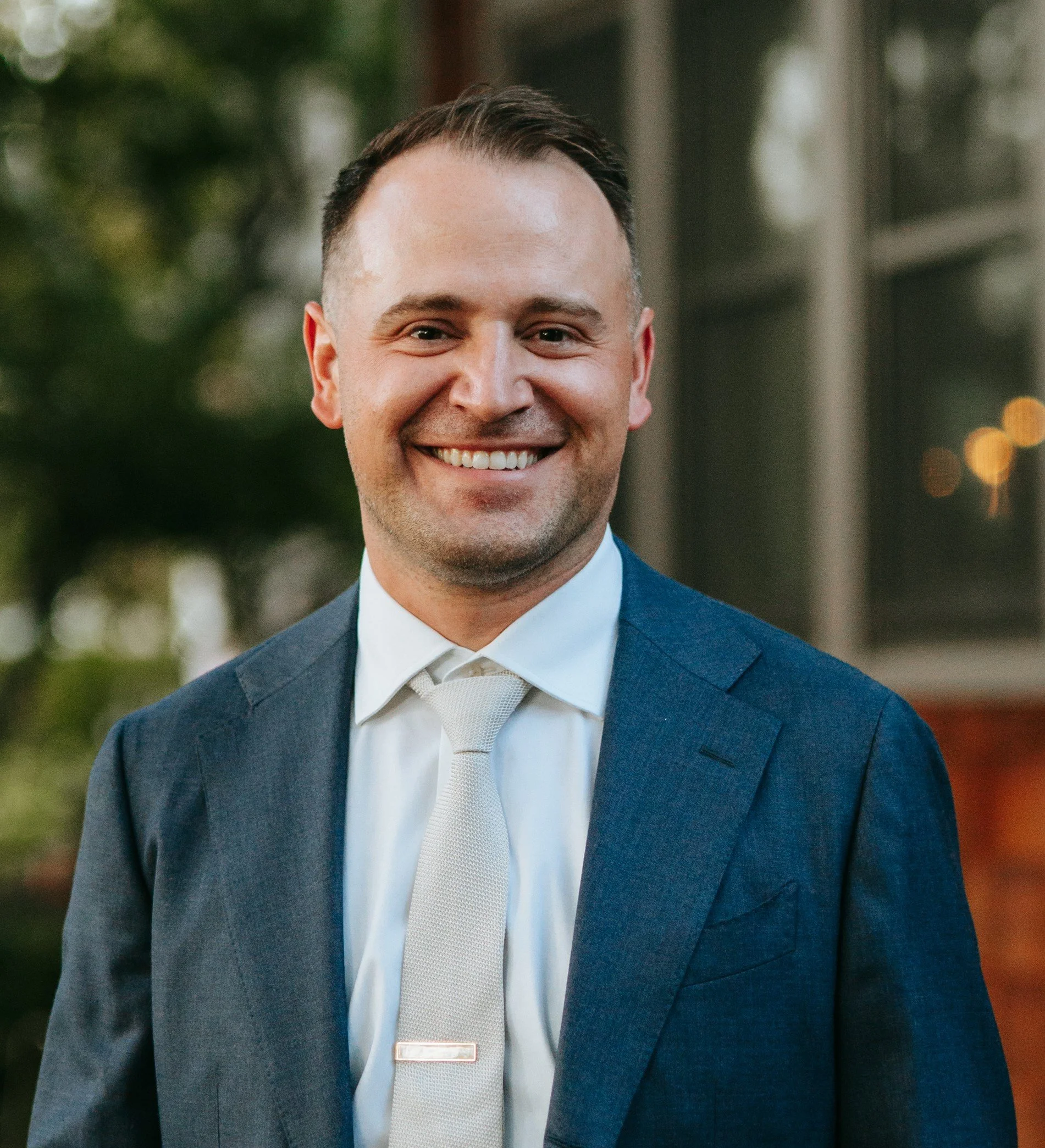 Joel Zink a man dressed in a blue suit, white shirt, and light-colored tie, smiling outdoors in front of a building with windows and greenery.