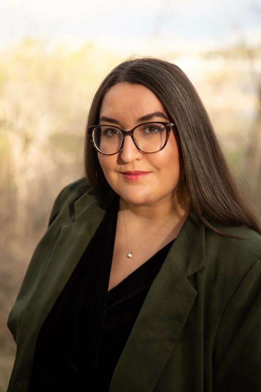 Karen DeAguero sits with long dark hair, glasses, and light skin, wearing a black top, olive green blazer, and a gold necklace, outdoors with a blurred natural background.