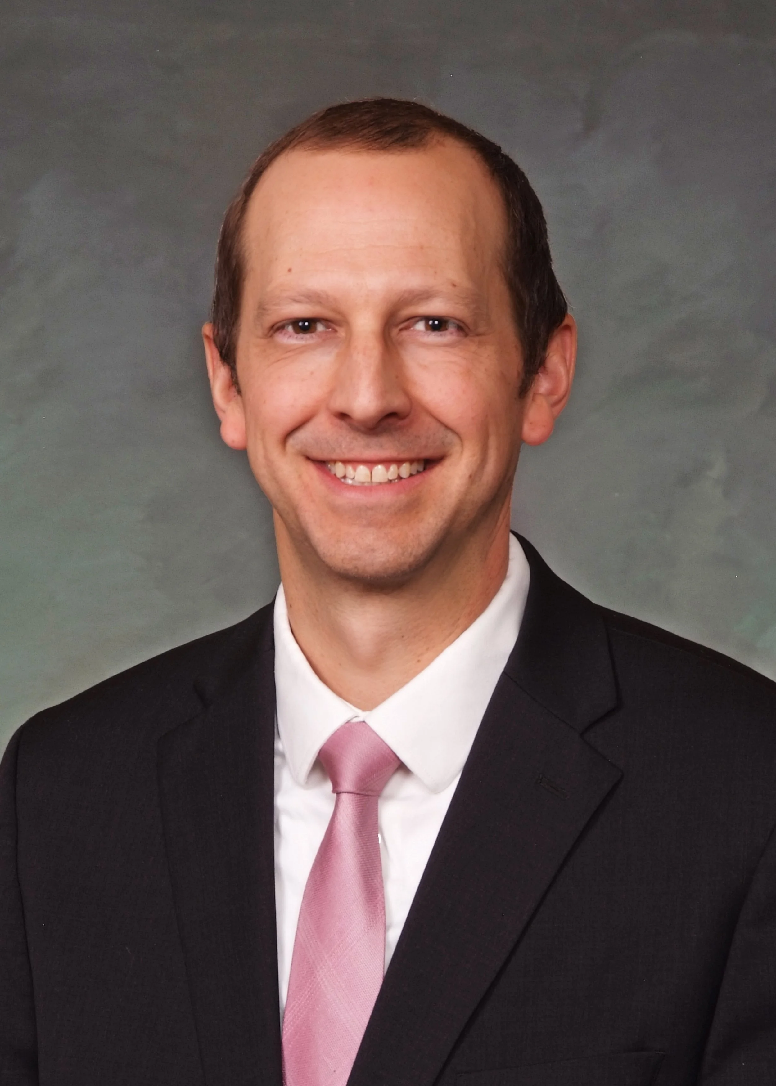 Andy in a professional portrait of a smiling man in a black suit, white shirt, and pink tie, against a mottled grey background.