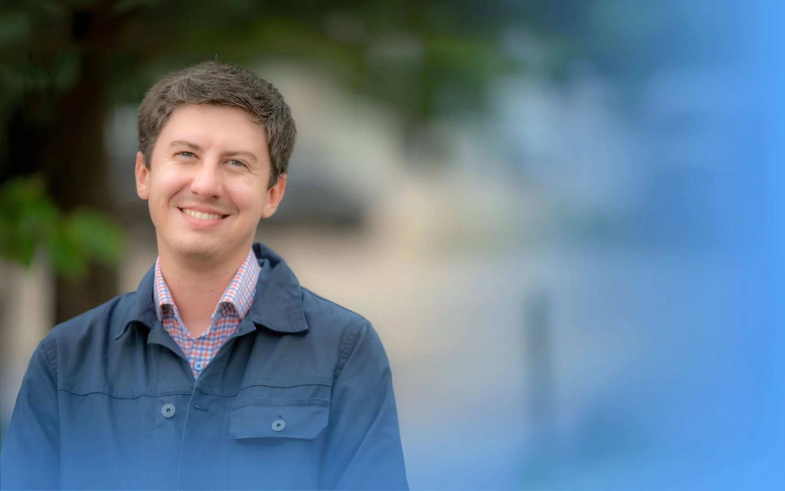 A smiling man (William) with short dark hair, wearing a checked shirt and a dark jacket, standing outdoors with a blurred background.