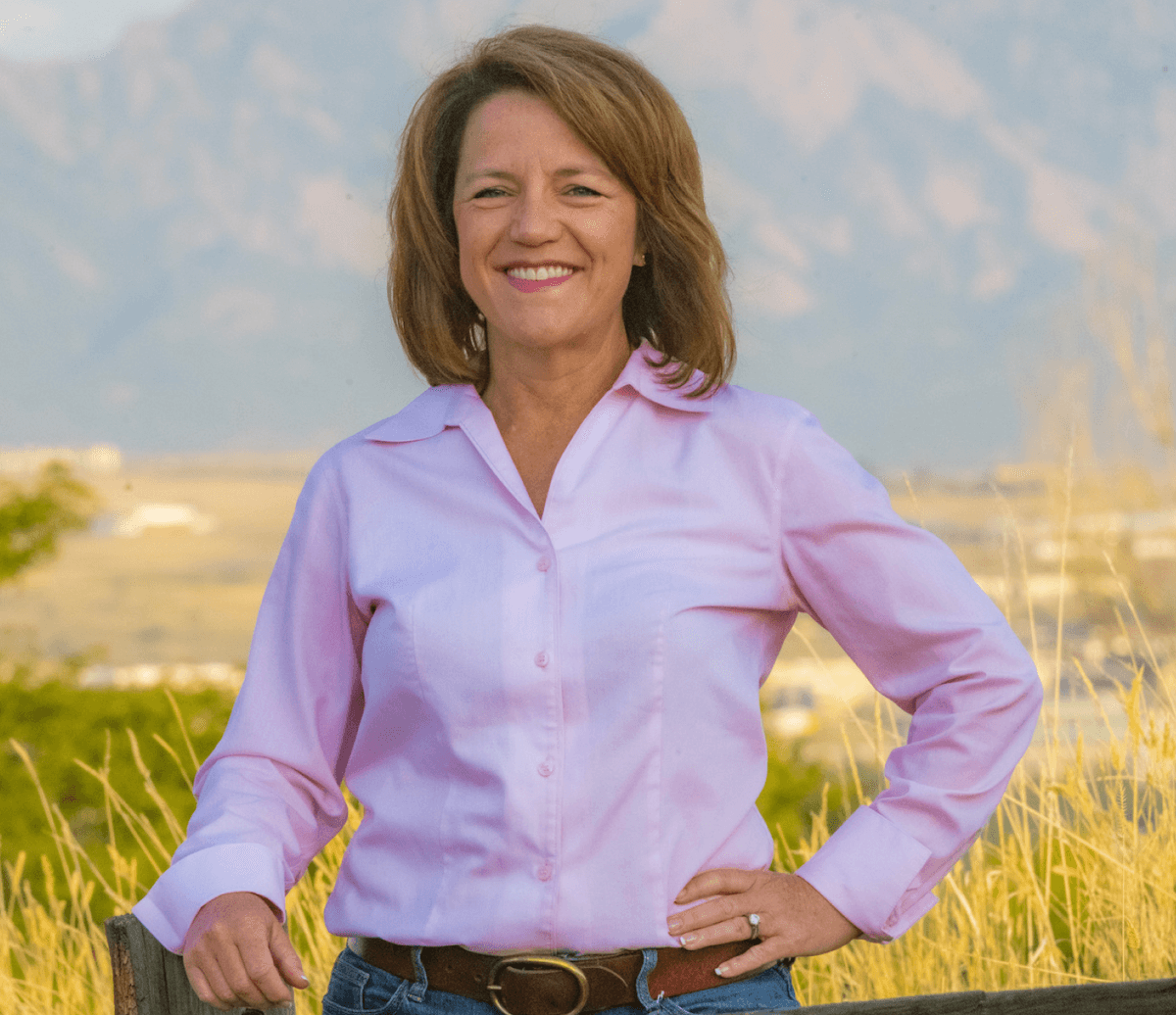Shannon Bird, a woman in a light pink button-up shirt standing outdoors, smiling, with golden fields and mountains in the background.