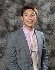 Austin ward sits with brown hair and a bright smile, wearing a light gray checkered blazer over a pink shirt, standing against a gray mottled background.