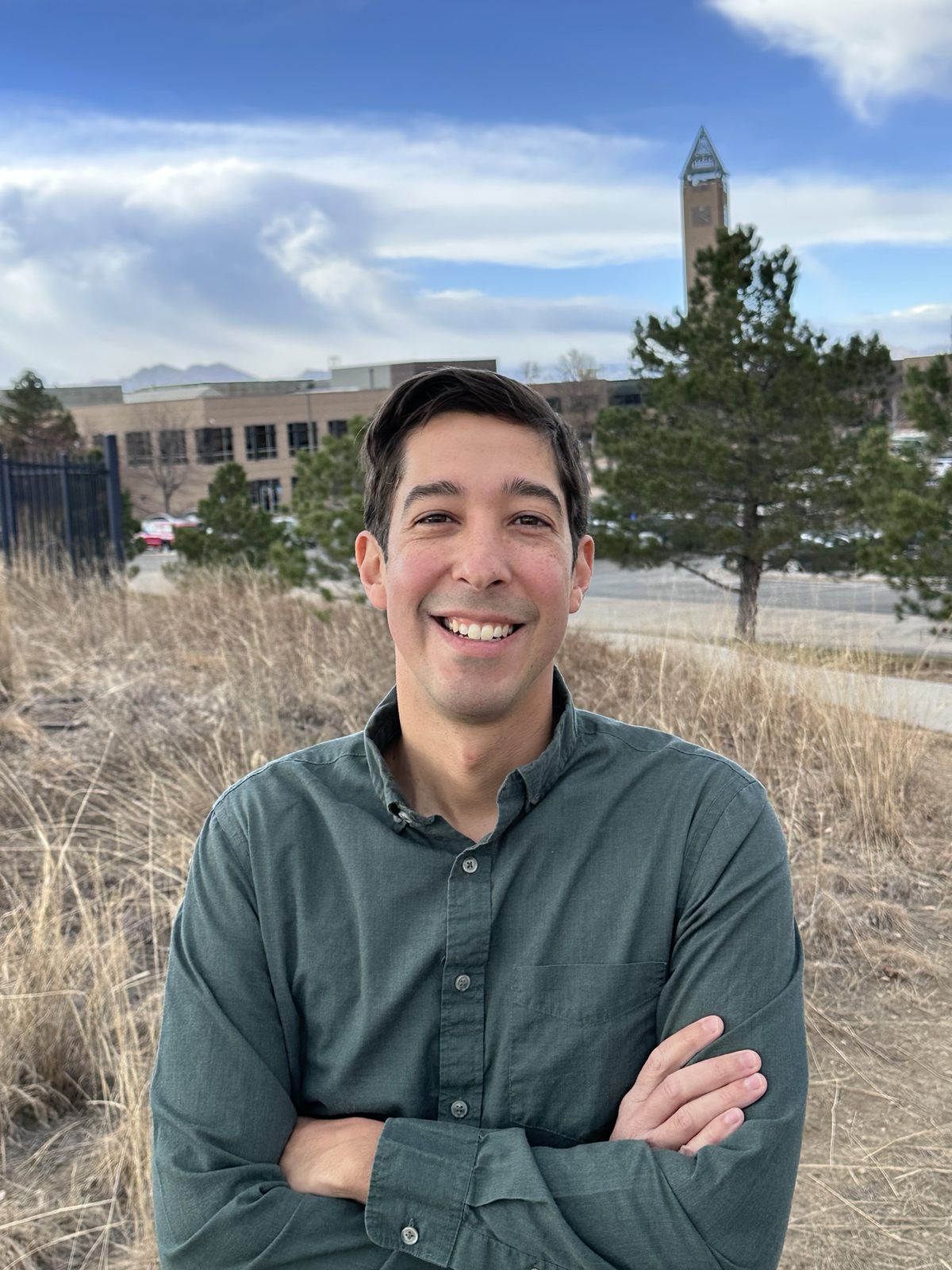 A young man Adam Barajas with dark hair smiling and crossing his arms outdoors, with dry grass, evergreen trees, and a clock tower in the background under partly cloudy skies.