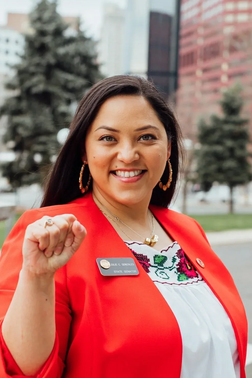 Julie, a woman with long dark hair smiling and making a fist gesture outdoors in front of trees and buildings. She is wearing a red blazer, a white embroidered blouse, large hoop earrings, and a name badge that reads 'Julie Gonzales, State Senator.'