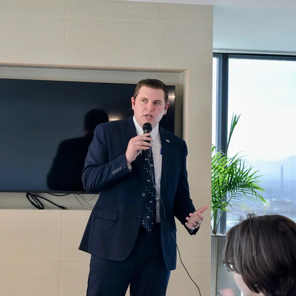 Michael in a navy suit speaking into a microphone in front of a large flat screen TV, with a window and a potted plant behind him in a modern conference room.