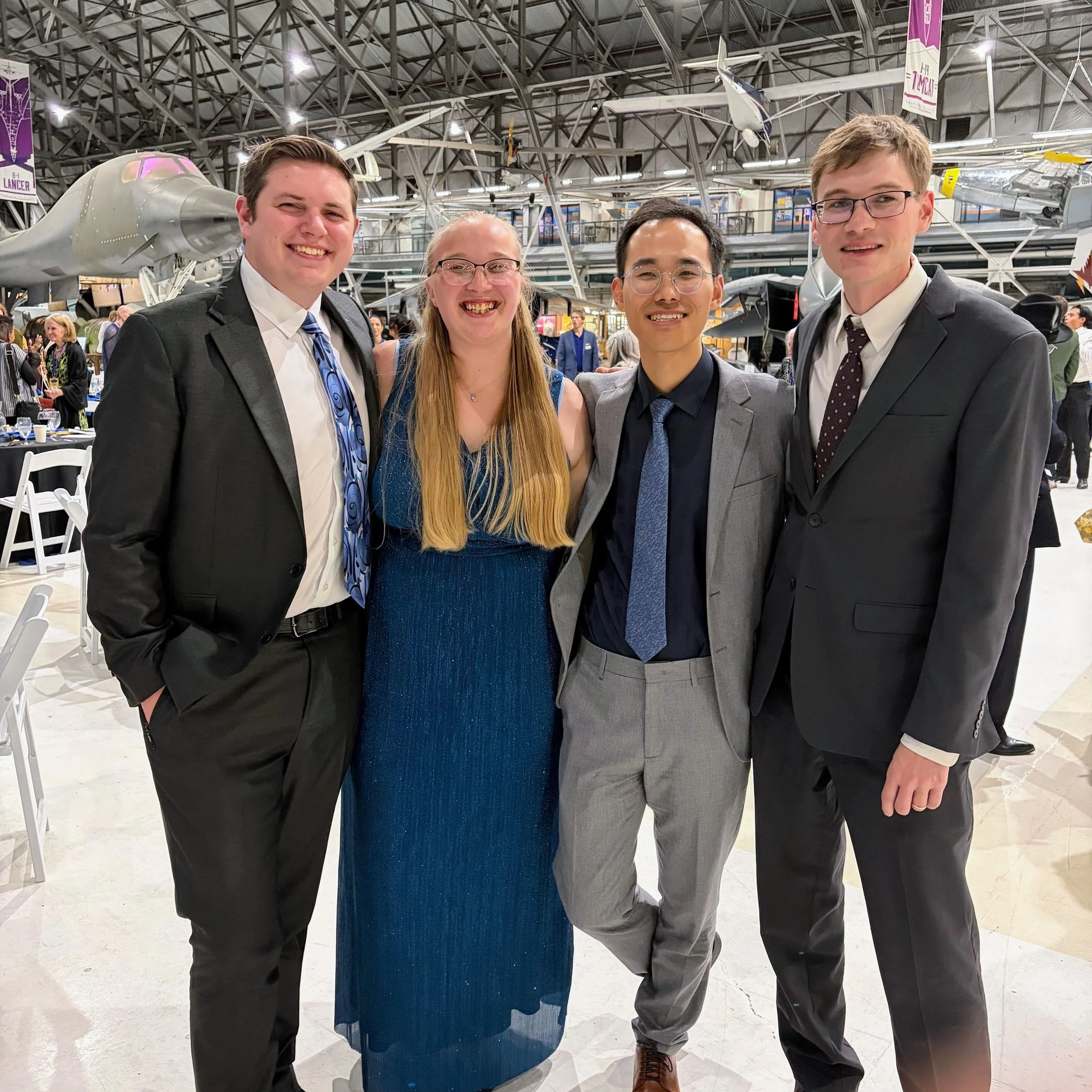 Four young adults, three men and one woman, dressed in formal attire, standing together at an indoor event, with aircraft exhibits and other attendees in the background.