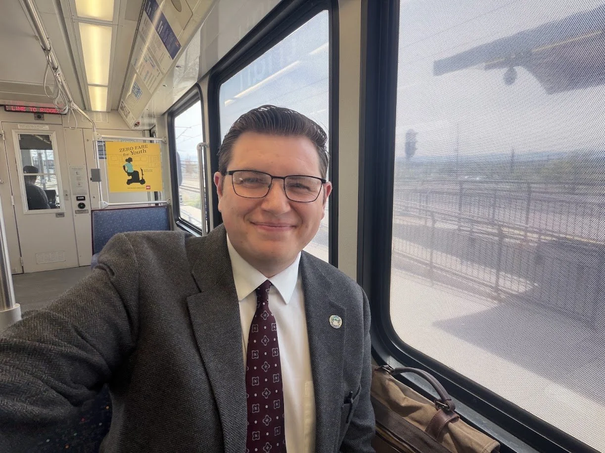 Smiling man in glasses and a suit taking a selfie on a train, with large windows showing the outside landscape.