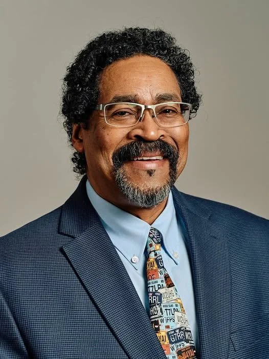 Mayor Steve Douglas, with curly black hair and glasses, wearing a blue suit and a colorful tie with license plates, smiling at the camera.