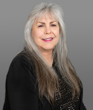 Portrait of an Kristi Douglas an older woman with long gray hair, smiling, wearing a black jacket and a dark top with small white dots.