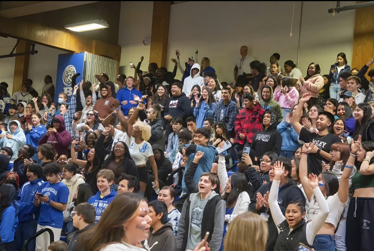 Large crowd of students and people in a gymnasium or auditorium, many smiling, cheering, or raising their hands, with some taking photos.
