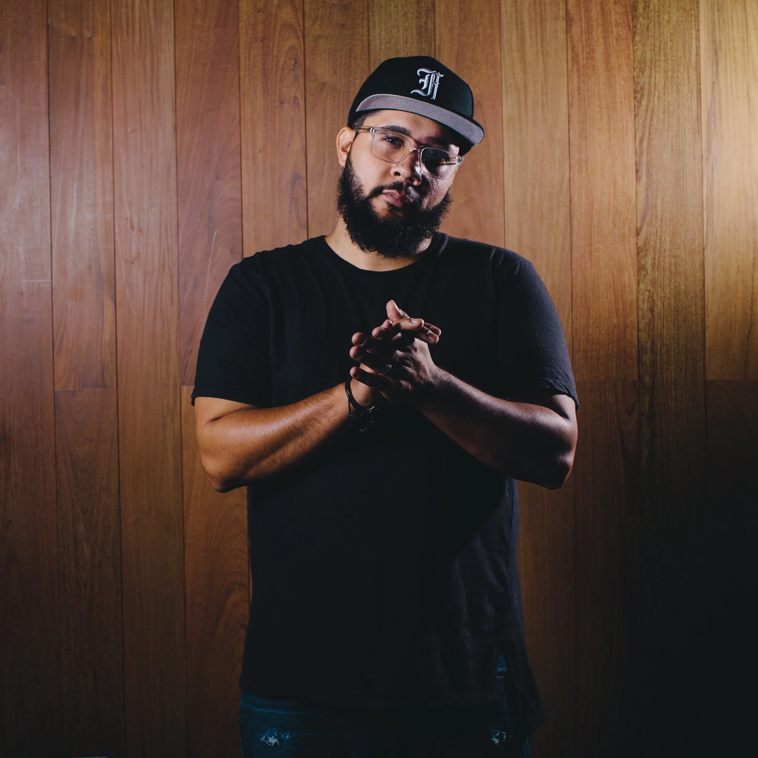 Music producer Ray Rock wearing a black hat stands in front of a wood-paneled wall with hands clasped, representing mentorship and creativity.