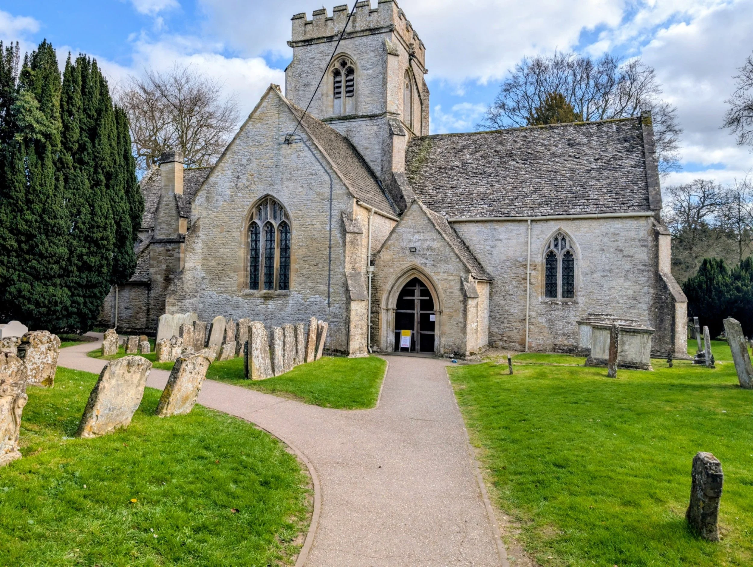 Ancient stone church with tall tower and arched windows, surrounded by graveyard and trees, under partly cloudy sky.