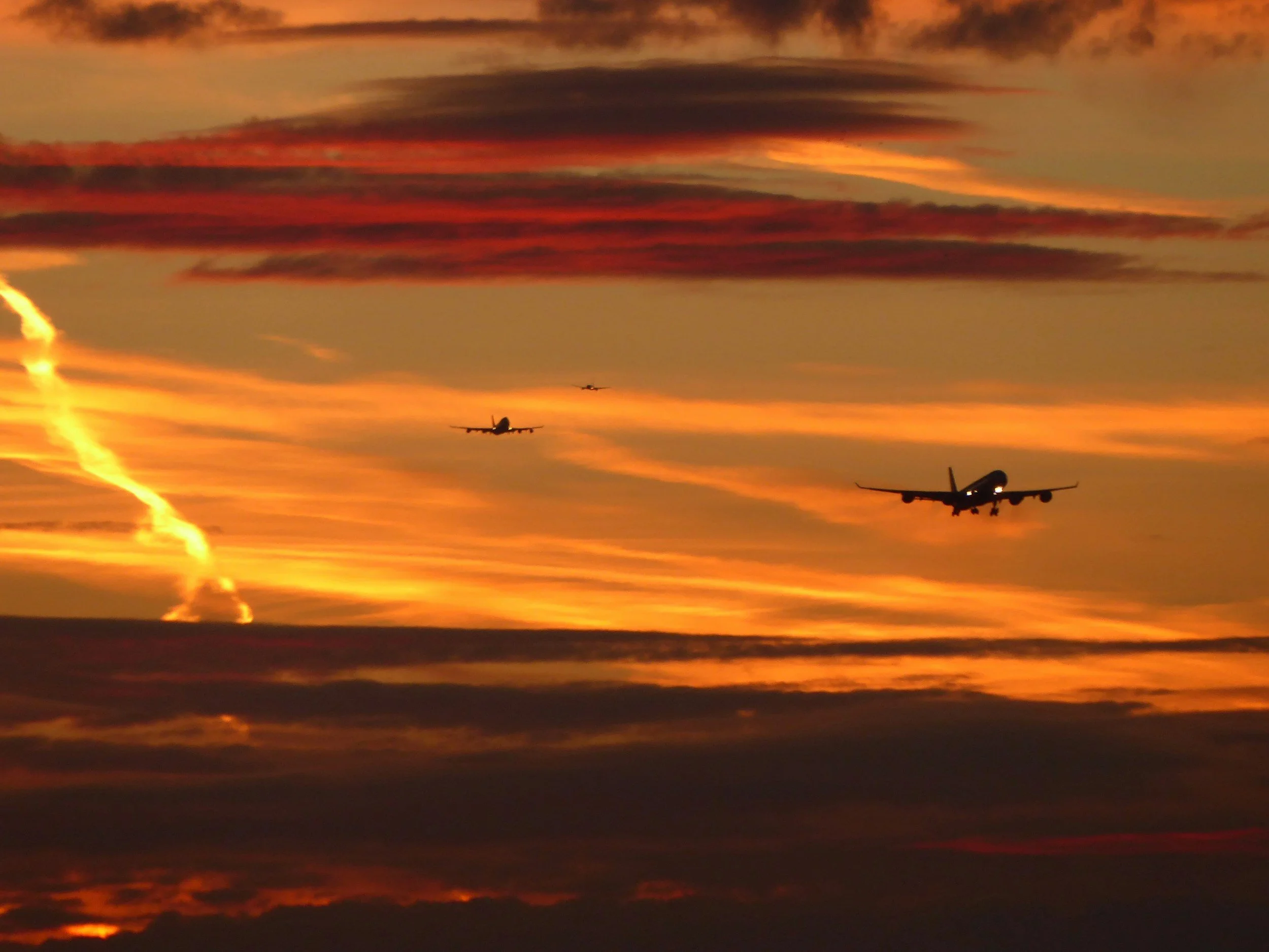 Silhouettes of three airplanes flying in front of a colorful sunset sky with orange, yellow, and dark clouds.