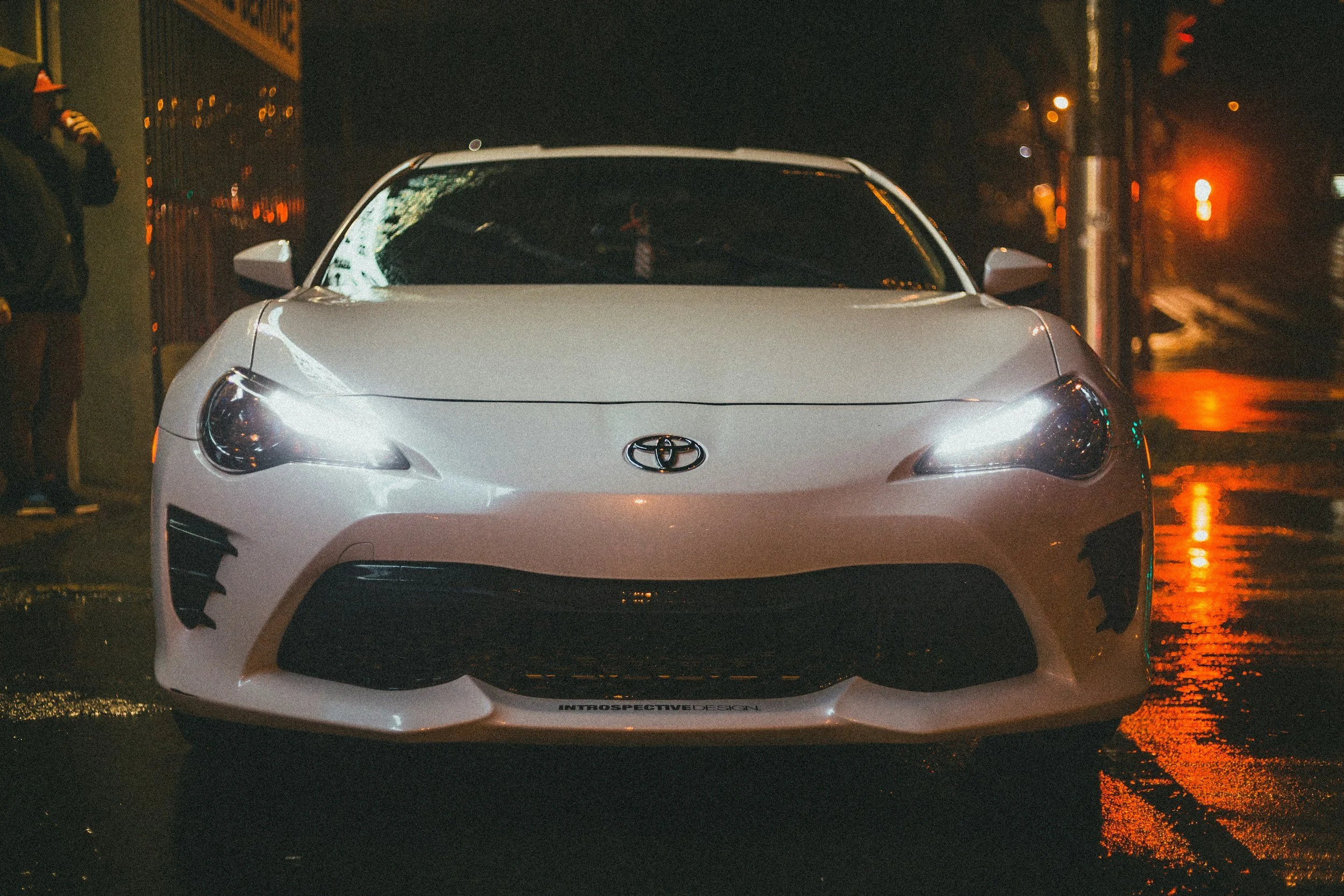 A white Toyota sports car parked on a wet street at night, with its headlights on and reflections visible on the road.