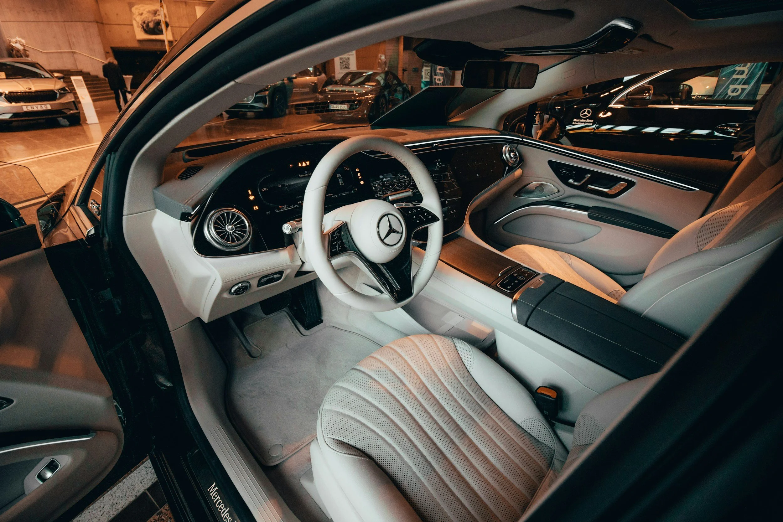 Interior of a luxury Mercedes-Benz car, showing the white leather seats, steering wheel, dashboard, and center console, parked indoors at a car show.