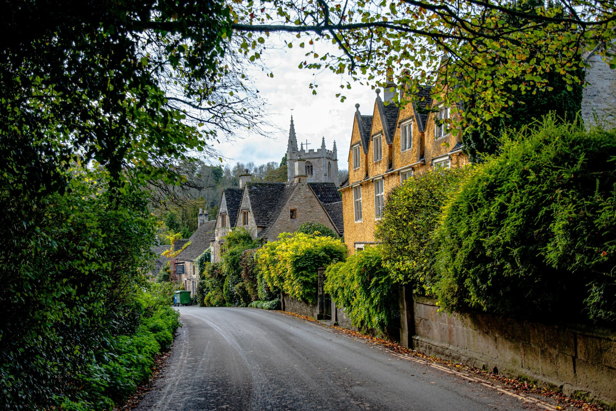 A winding residential street in a small town with lush greenery, stone houses, and a church steeple in the background.