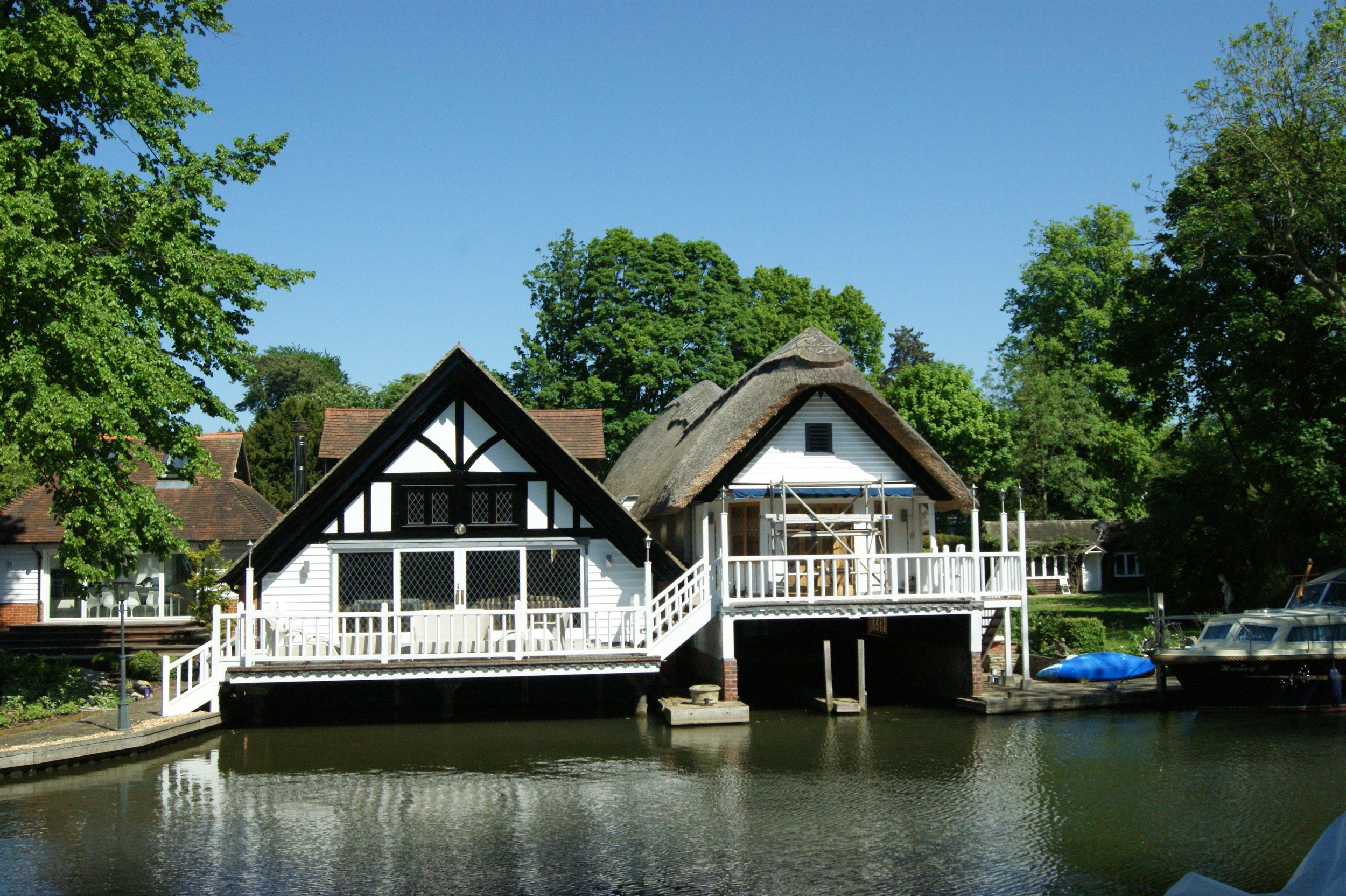 A waterfront house with a thatched roof and black-and-white half-timbered design, surrounded by green trees under a clear blue sky, with boats docked along the water.