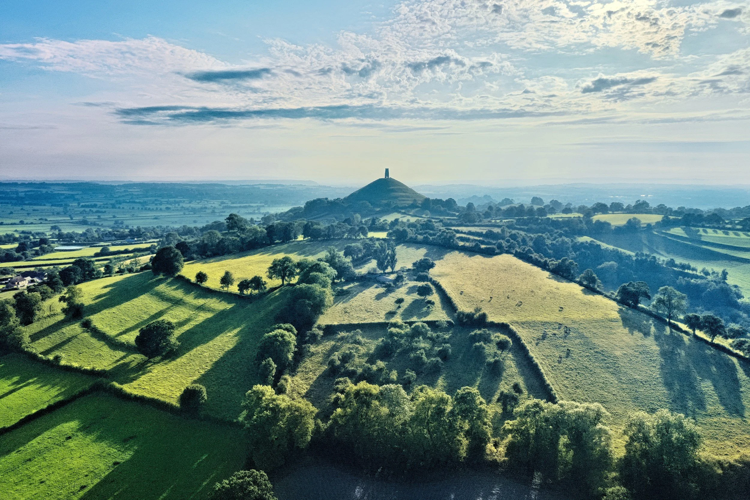 A scenic aerial view of rolling green hills with trees and patchwork fields, with a hill featuring a tower on top in the distance under a partly cloudy sky.