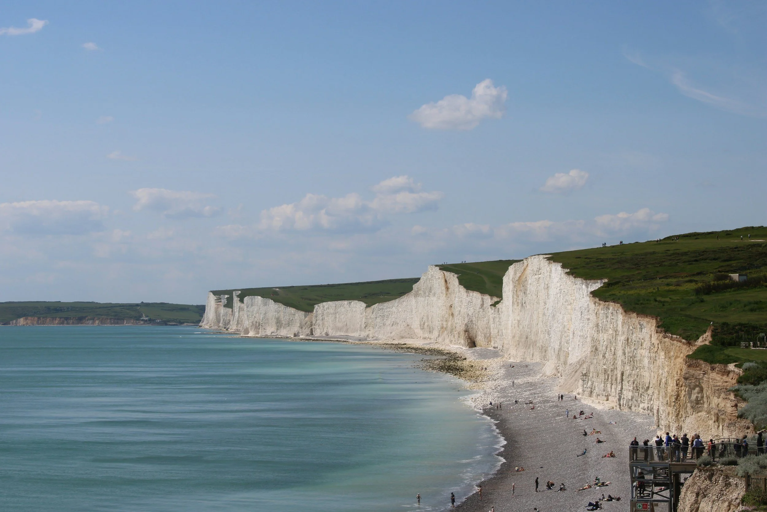 White chalk cliffs along a pebble beach with people walking and sitting, under a partly cloudy sky.
