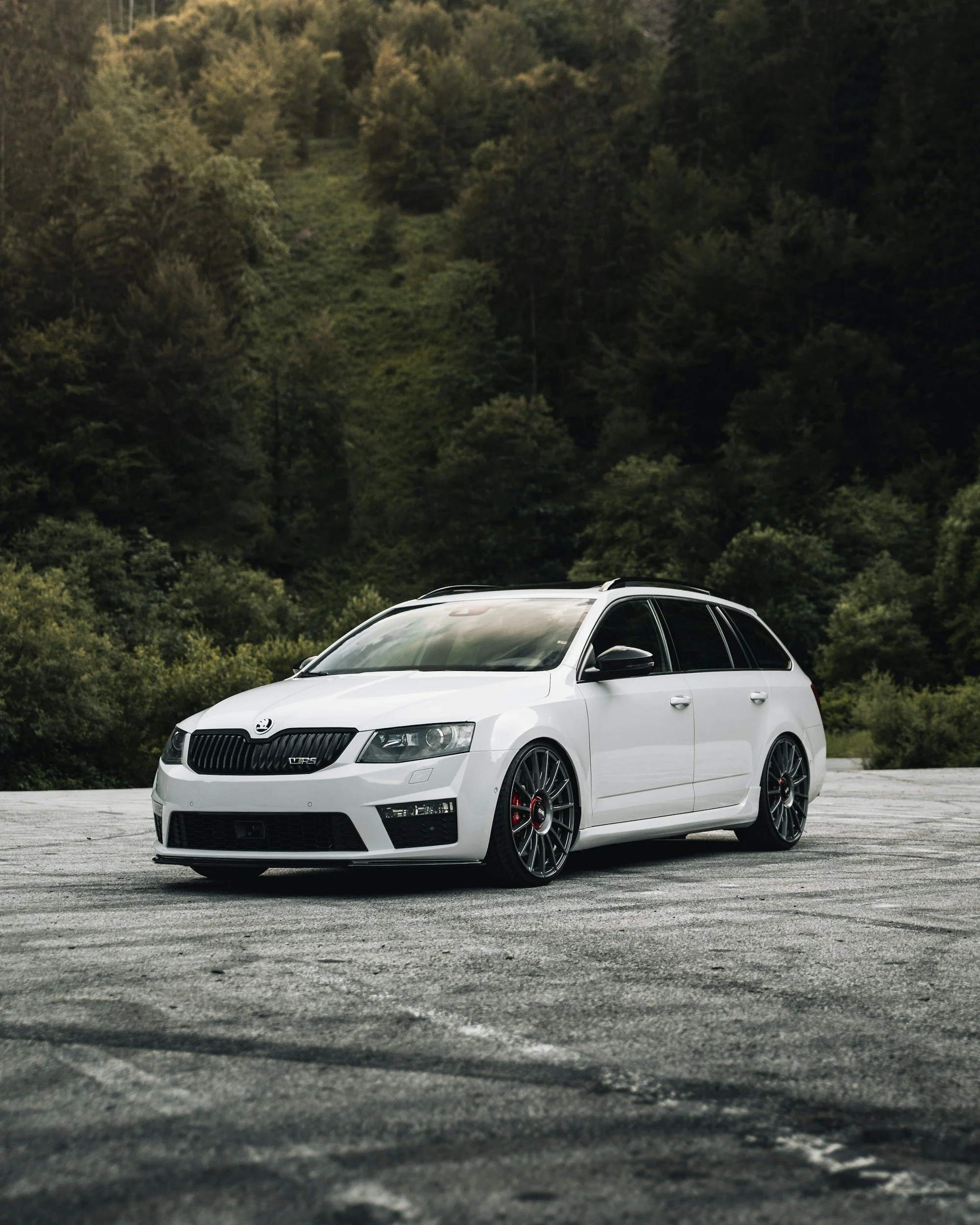 A white station wagon parked on a paved area with a forested hillside in the background.