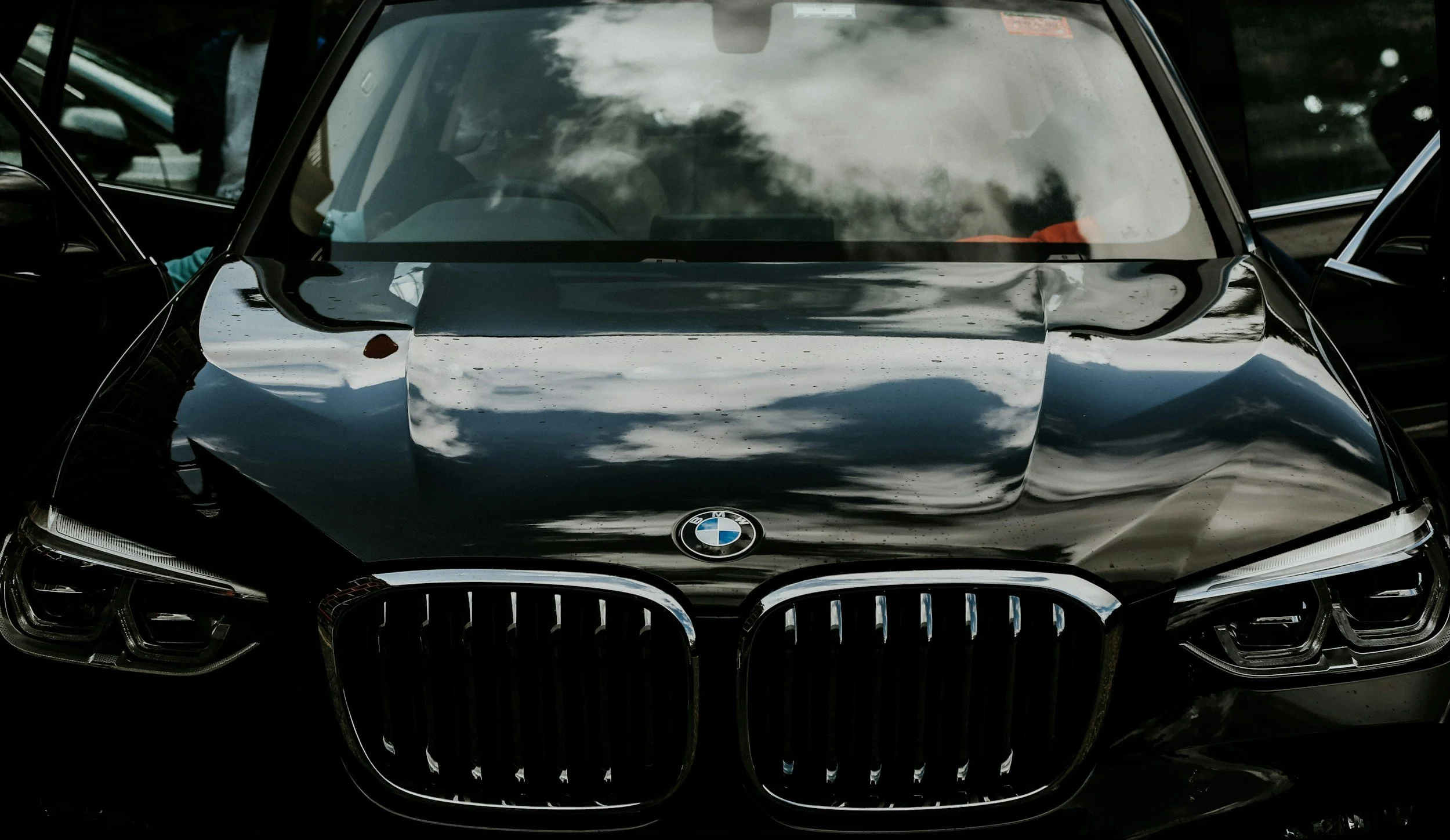 Front view of a black BMW car with a shiny, reflective surface, showing the grille, headlights, and windshield, with reflections of clouds on the hood and windshield.