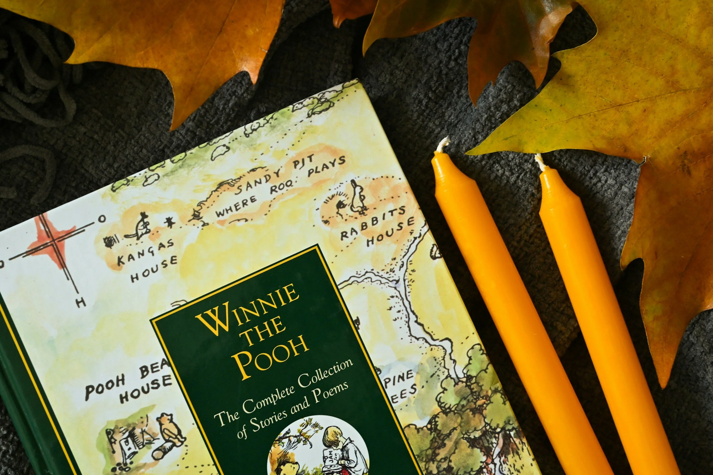 Copy of Winnie the Pooh book, two yellow beeswax candles, and autumn leaves on a dark fabric surface.