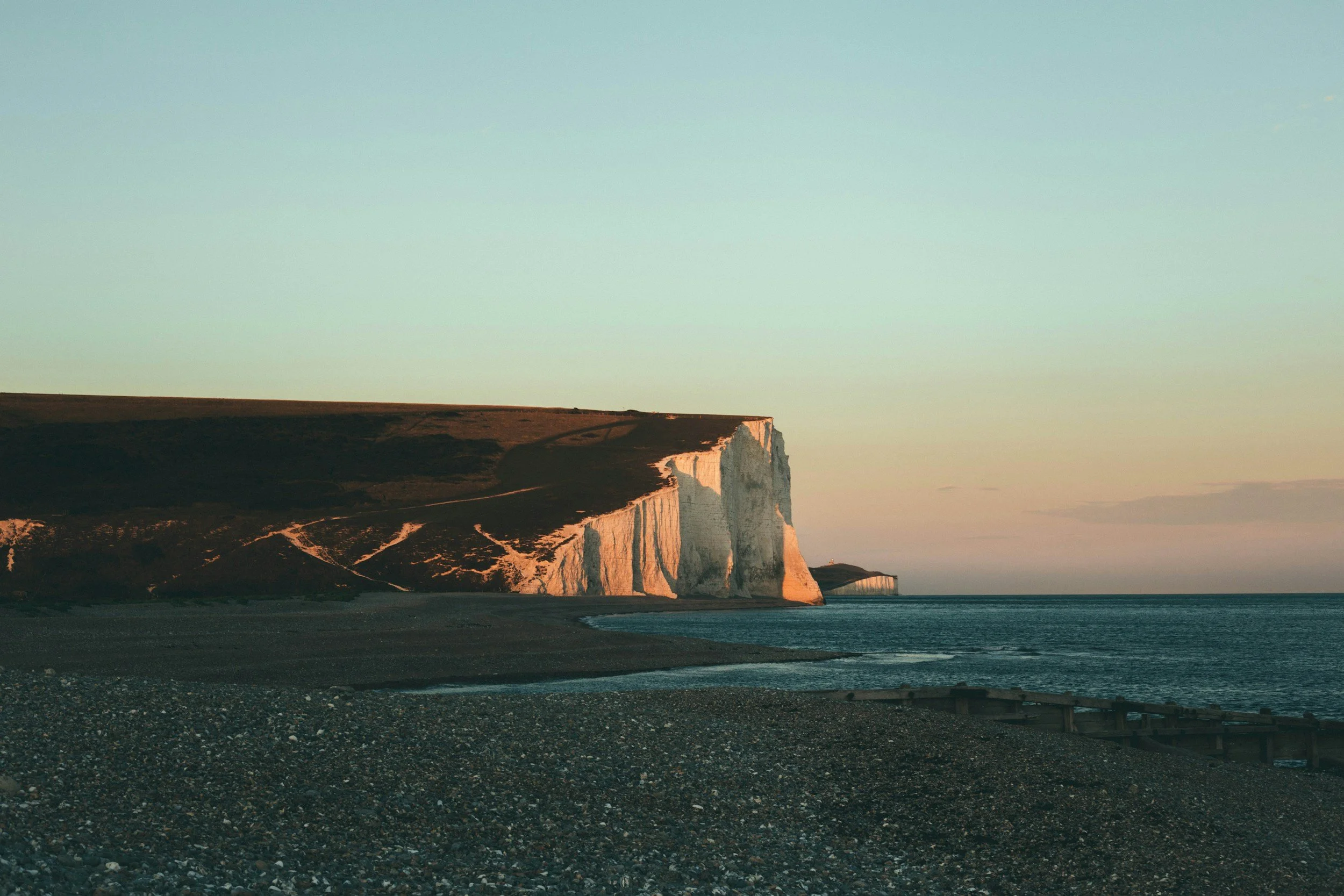 Cliffs of white chalk rise above a rocky beach at sunset, with the sky transitioning from light blue to warm orange hues.