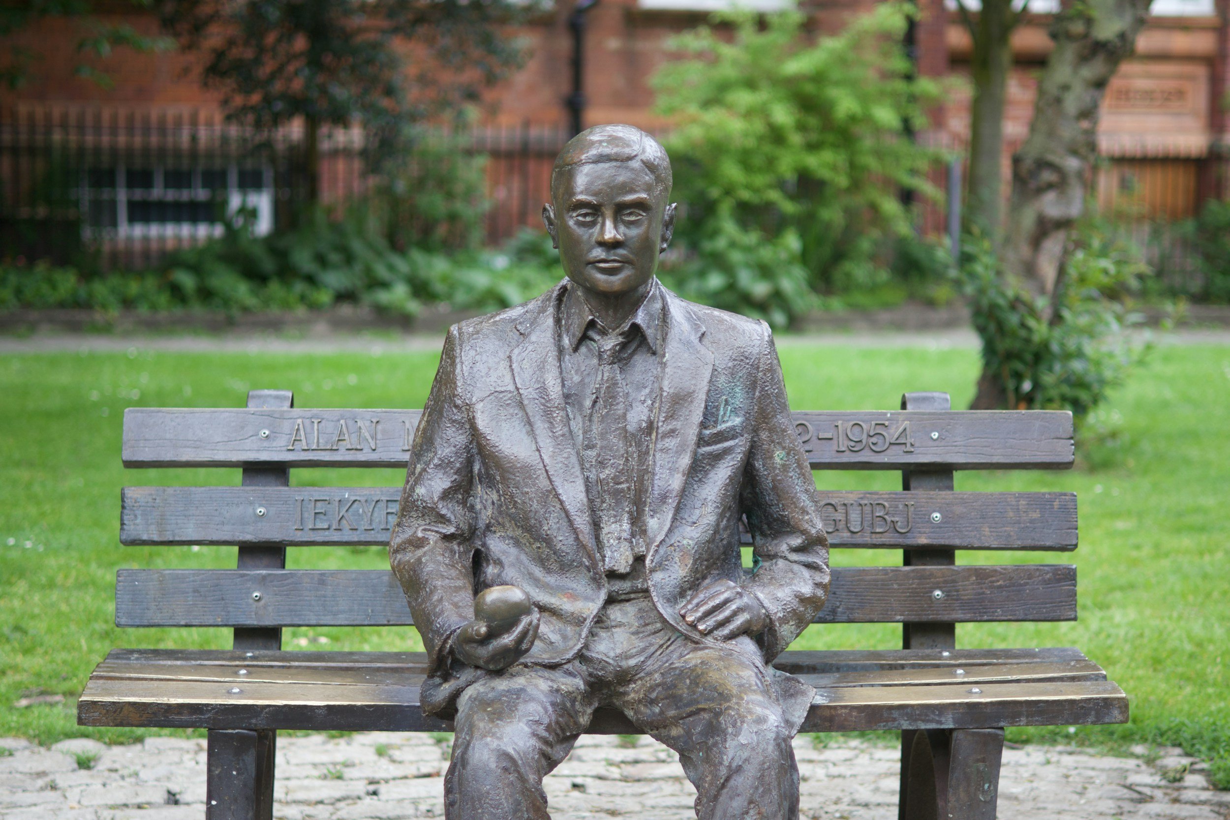 Bronze statue of a seated man on a park bench with greenery and brick wall in the background.