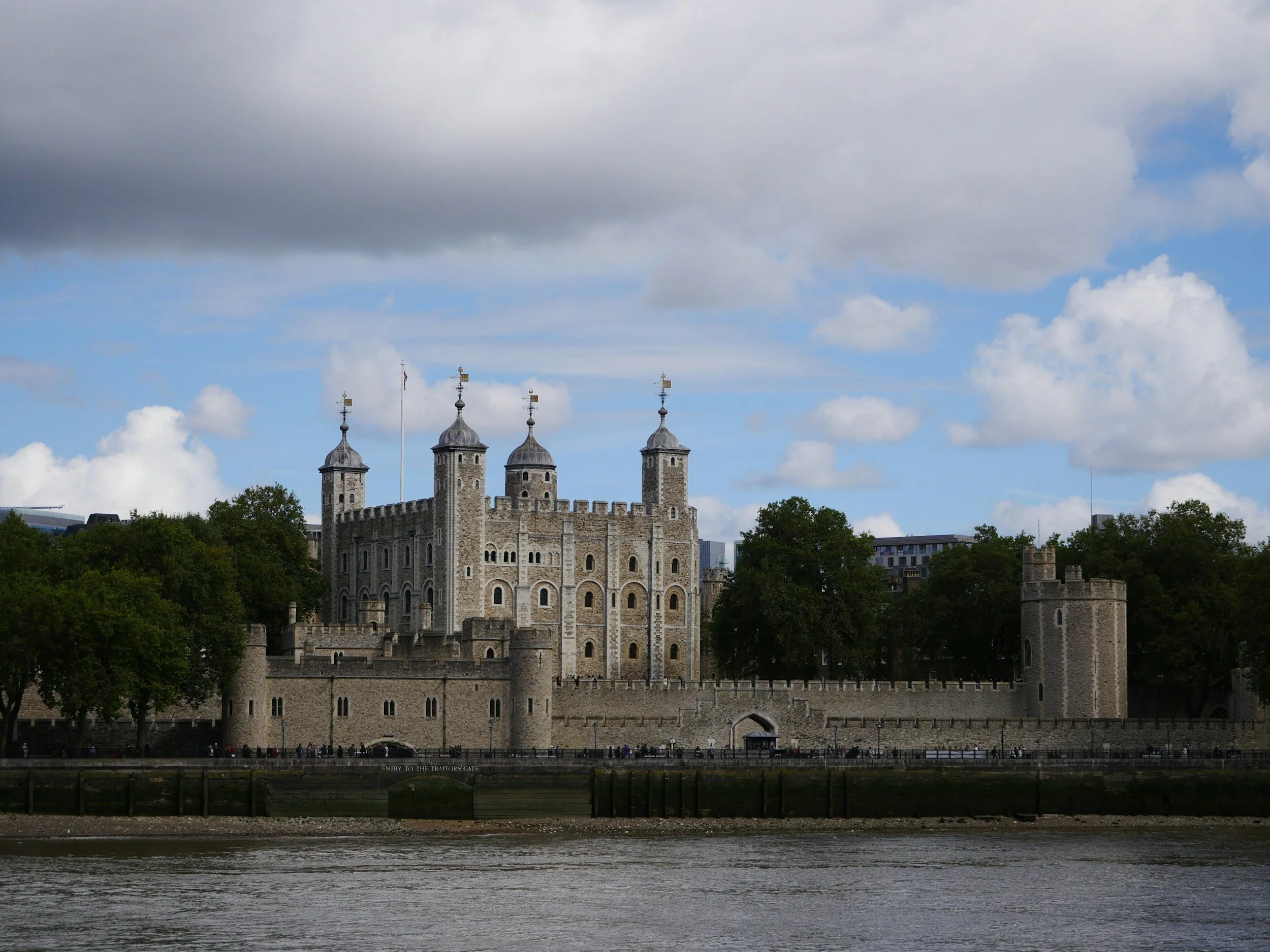 A historic stone castle with four towers is situated along a river, with a partly cloudy sky overhead and trees surrounding the castle.