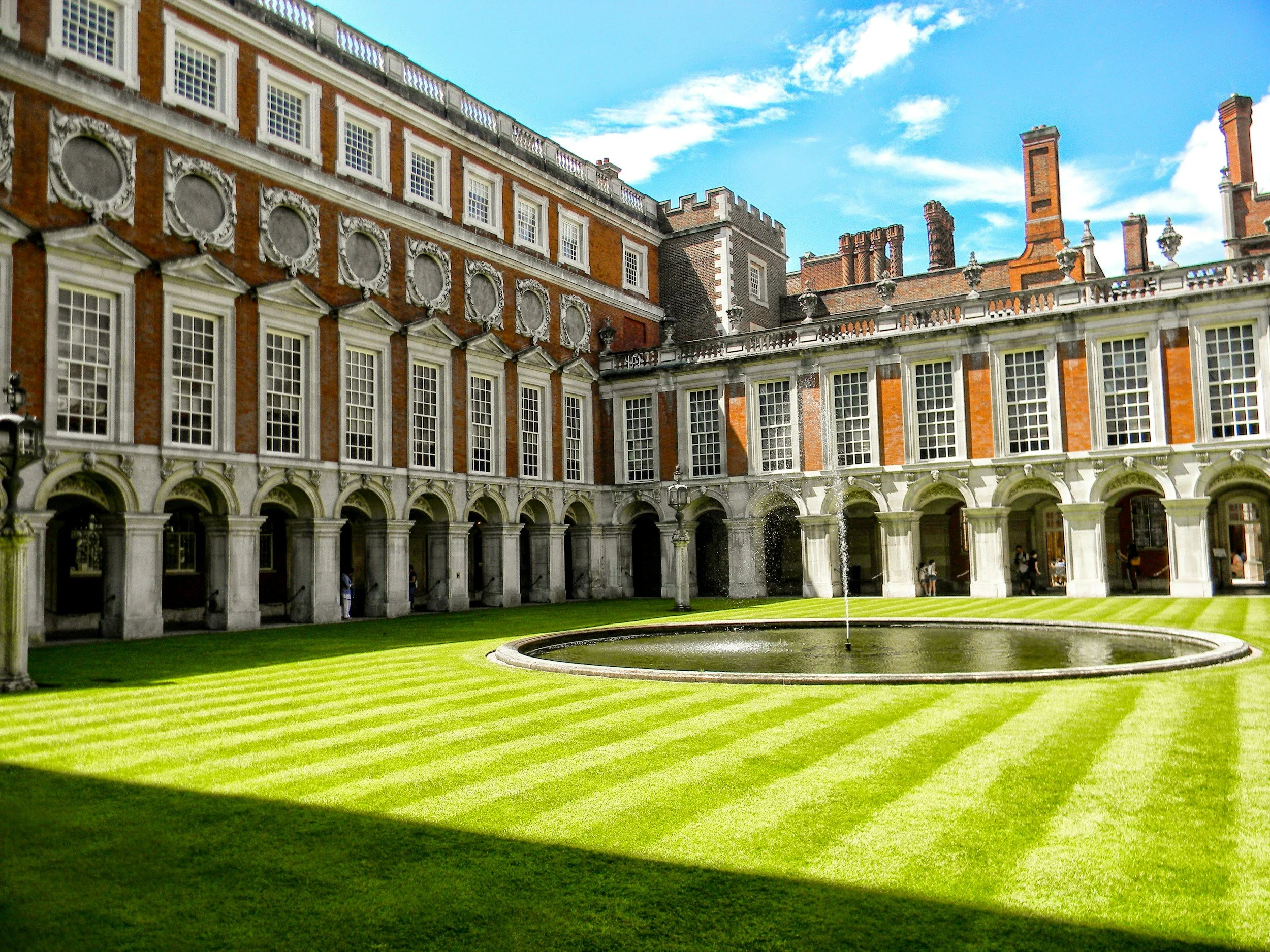 The image shows a large historic mansion with red brick exterior, white decorative trim, numerous windows and chimneys, and a courtyard with a fountain and green grass.