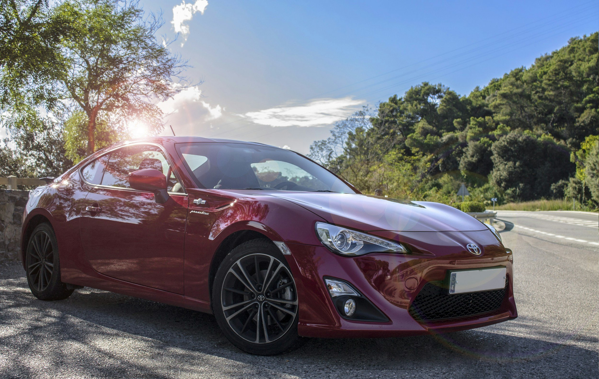 A red Toyota sports car parked on the side of a road next to trees and green foliage under a bright blue sky with some clouds, with the sun shining and flaring in the image.