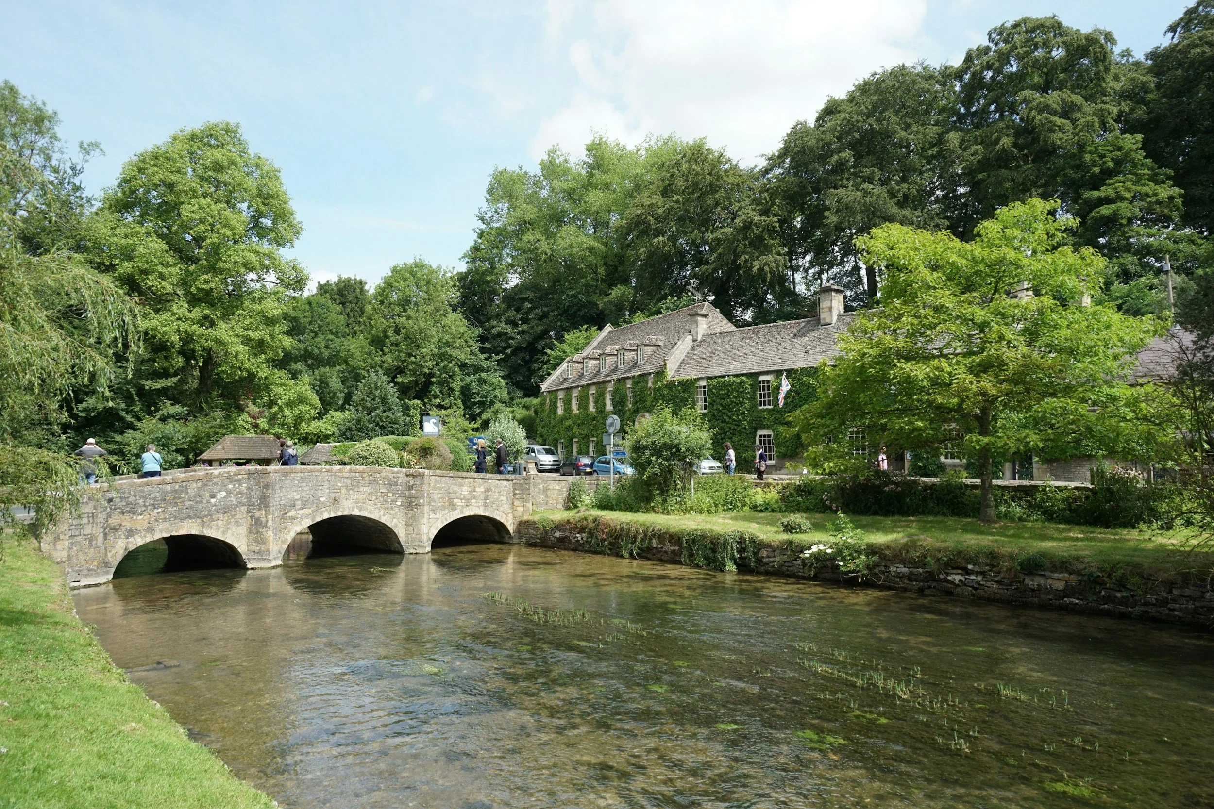 A scenic view of a stone bridge over a river with a historic building and lush green trees in the background on a bright, partly cloudy day.
