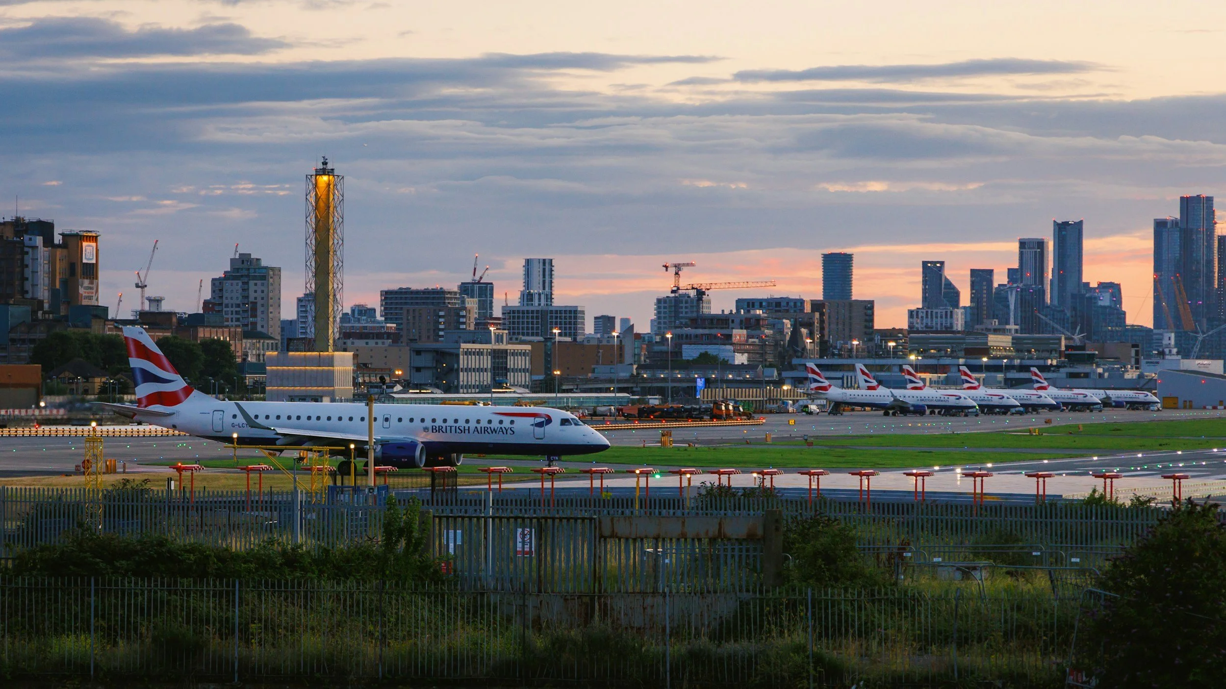 Skyline of a city at sunset with high-rise buildings, construction cranes, and British Airways airplanes parked on the tarmac of an airport in the foreground.