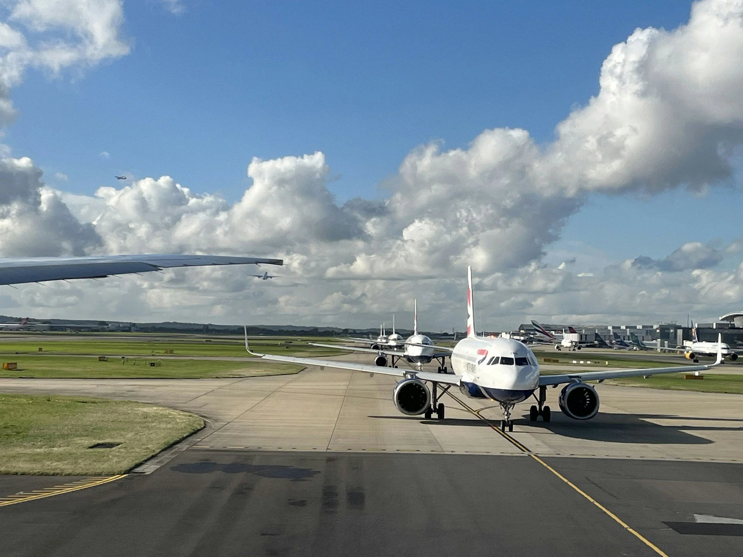 View of an airport tarmac with several airplanes parked, including a crowed airplane in the foreground, and other planes in the background. The sky is partly cloudy with some blue sky visible.