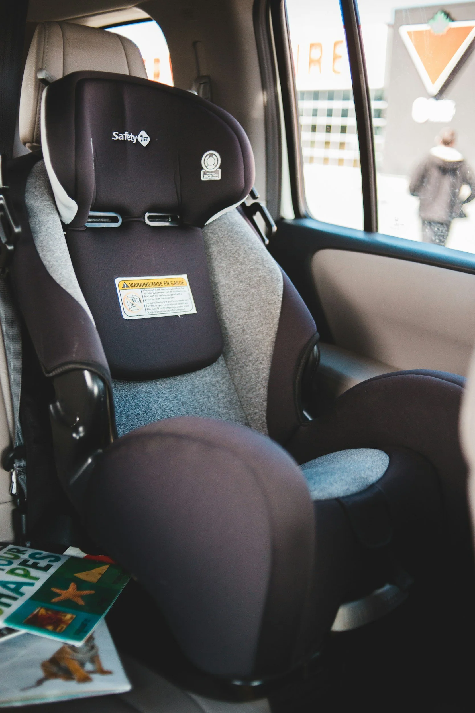 Inside a vehicle, showing a child safety car seat attached to a car seat belt, with a window and a person outside standing near a sign in the background.