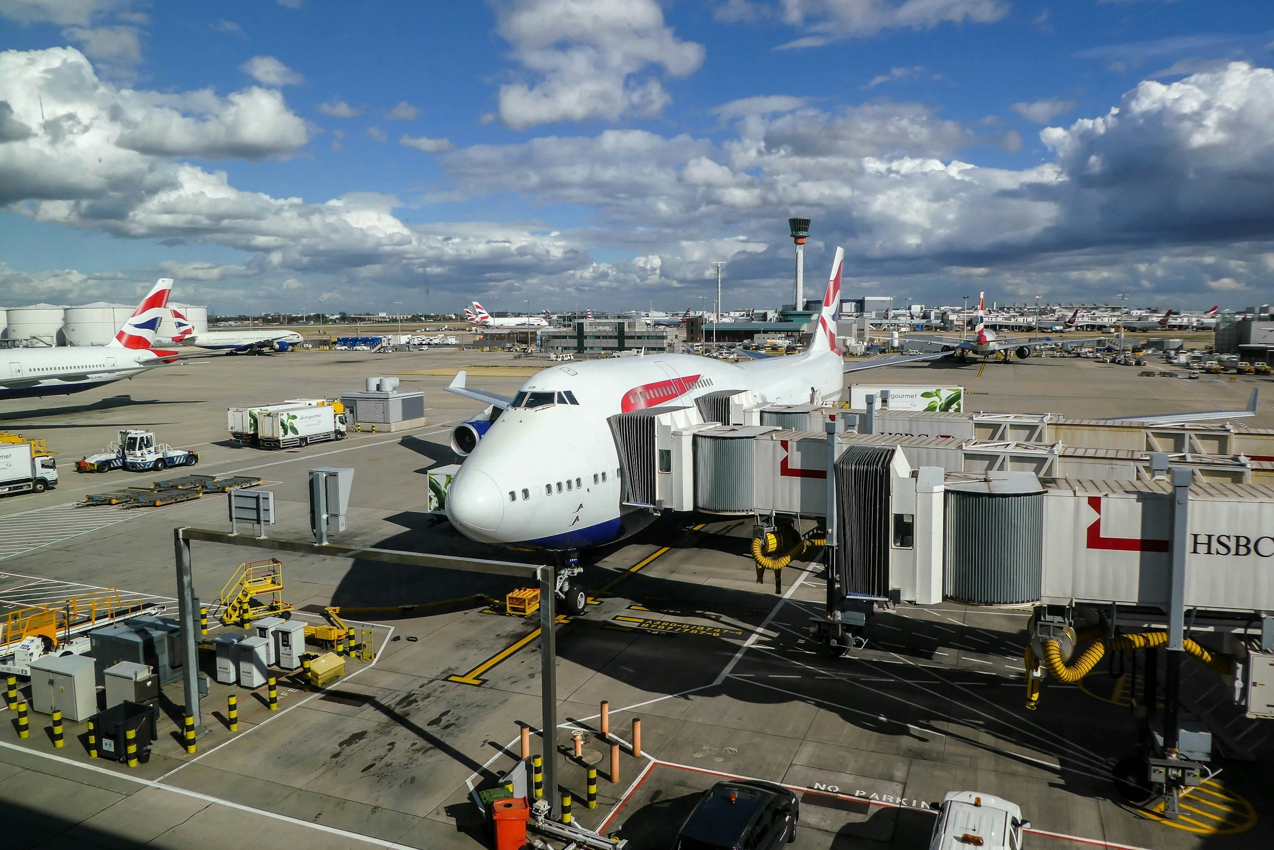 View of an airport tarmac with multiple parked airplanes, airport vehicles, and a control tower in the background under a partly cloudy sky.