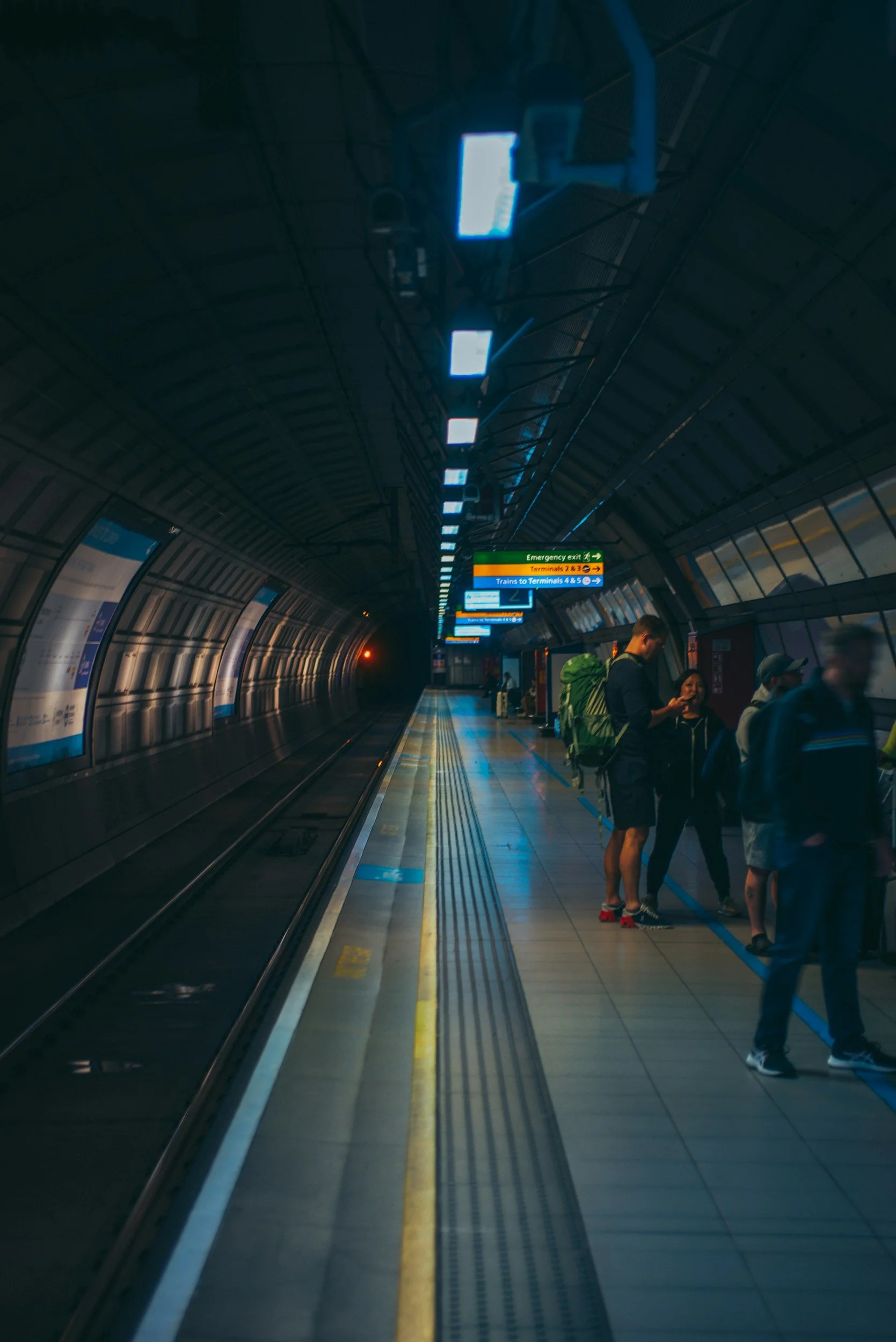 People waiting on an underground train platform with overhead signs showing directions to various terminals and trains.