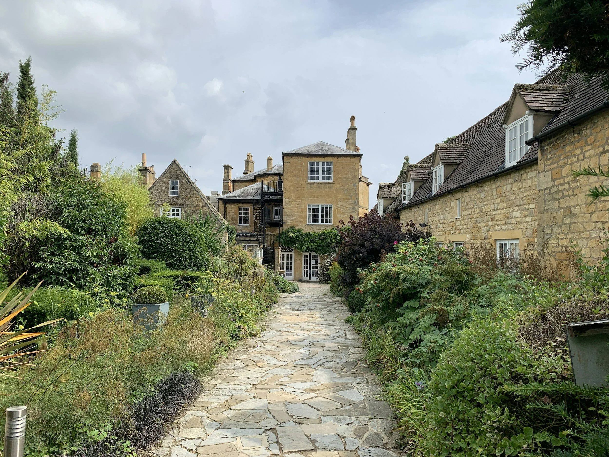 A stone pathway in a lush garden leading to charming houses with stone facades and pitched roofs, surrounded by greenery and flowering plants.