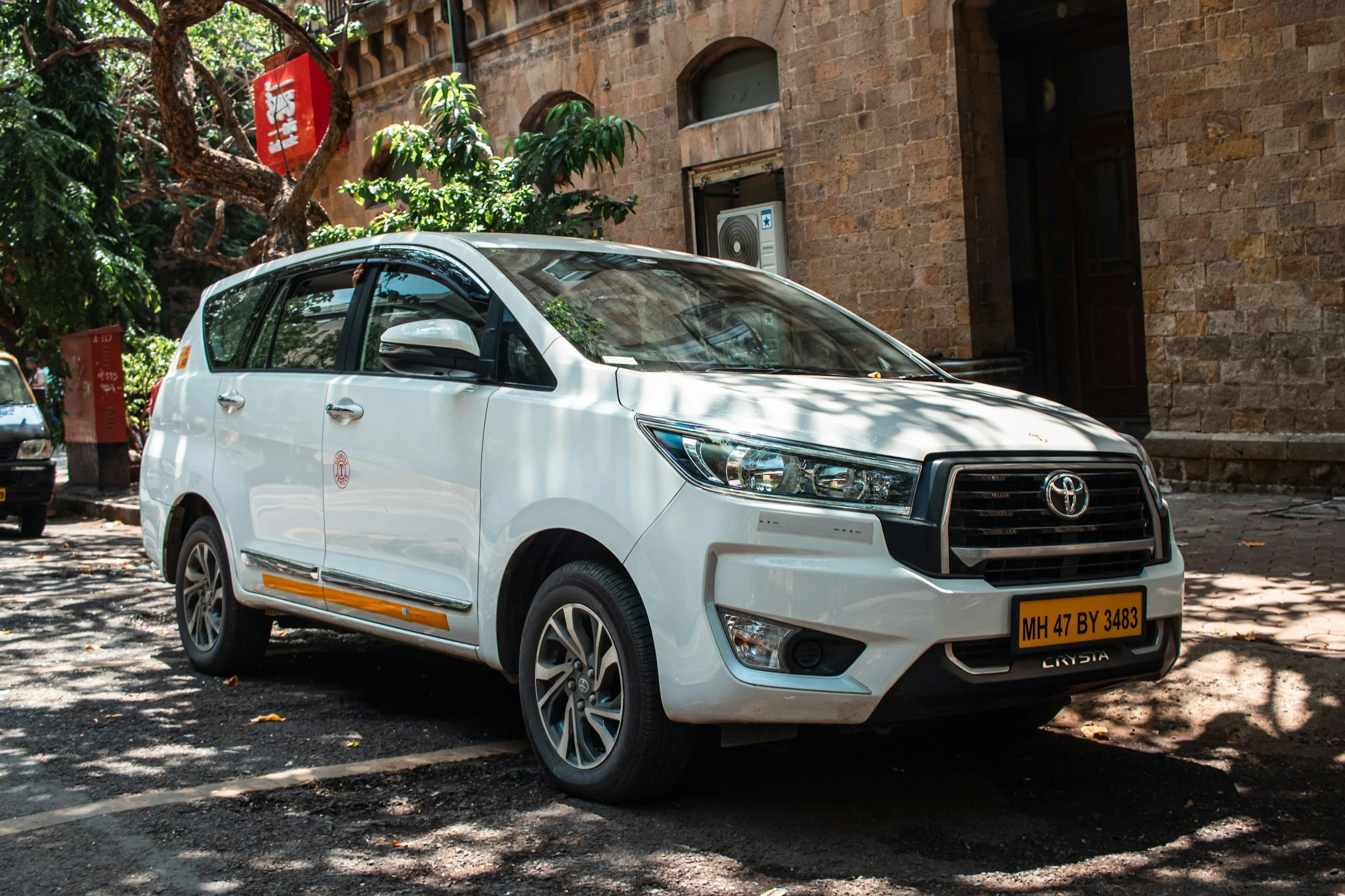 A white Toyota car with the license plate 'MH 47 BY 3483' parked on a tree-shaded street next to a brick building with arched windows and an air conditioning unit. Trees and leaves are visible above and around the car.