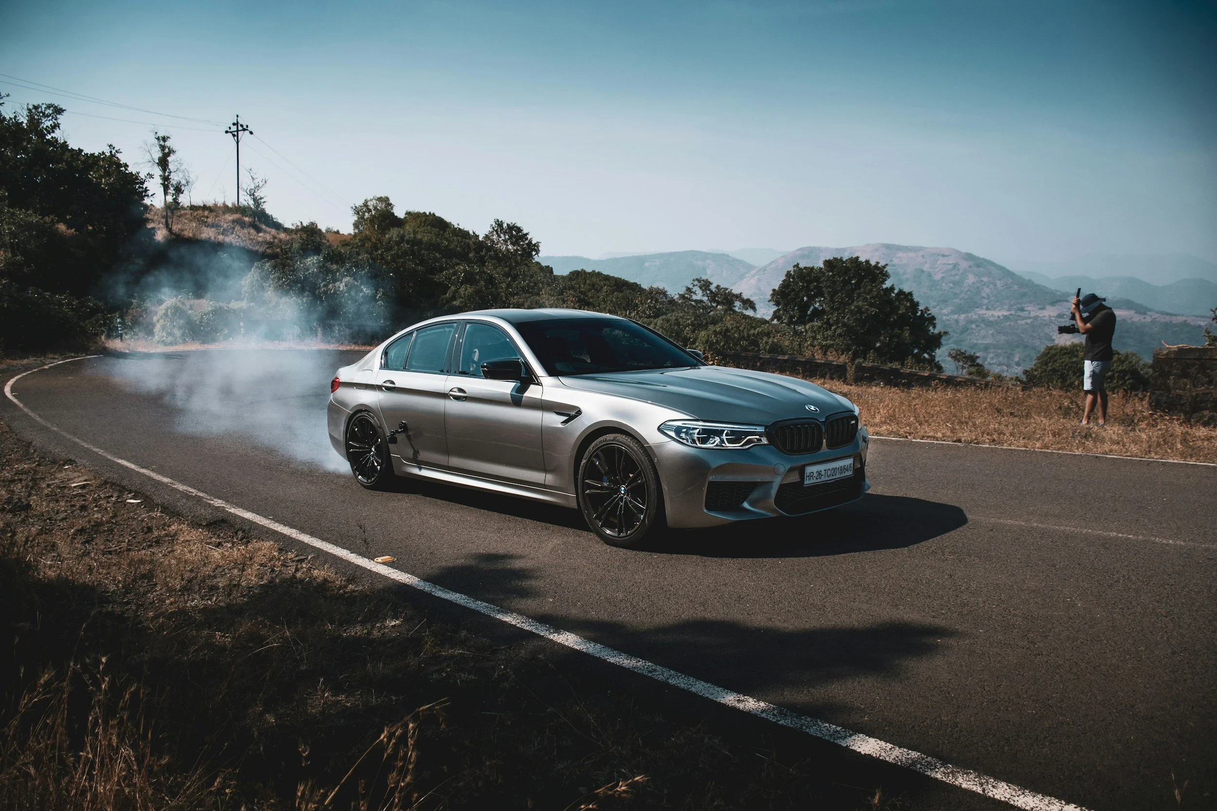 A silver BMW sedan driving on a winding mountain road with a person taking a photo nearby, surrounded by trees, mountains, and clear blue sky.