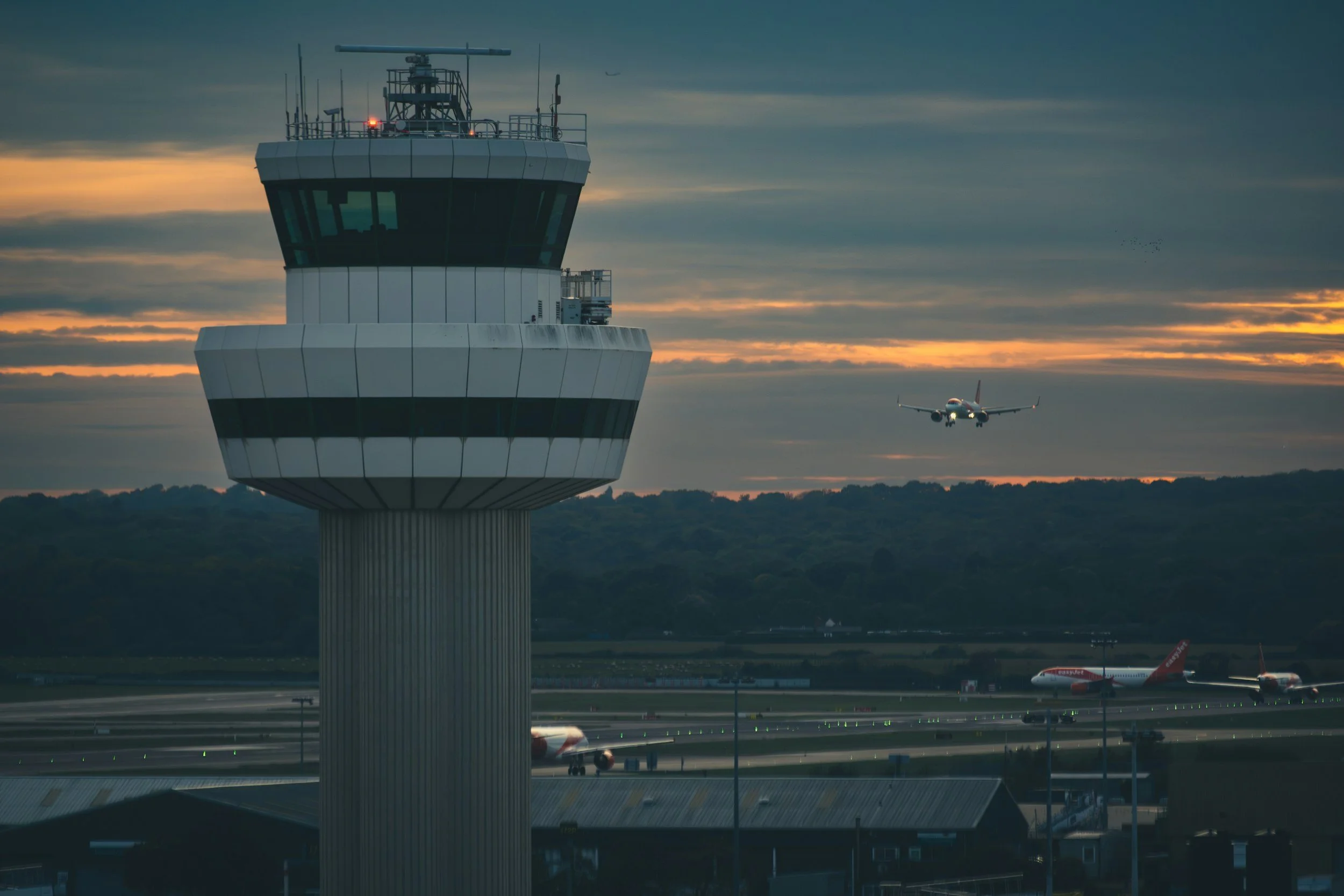 An airport control tower with a plane taking off in the background during sunset.
