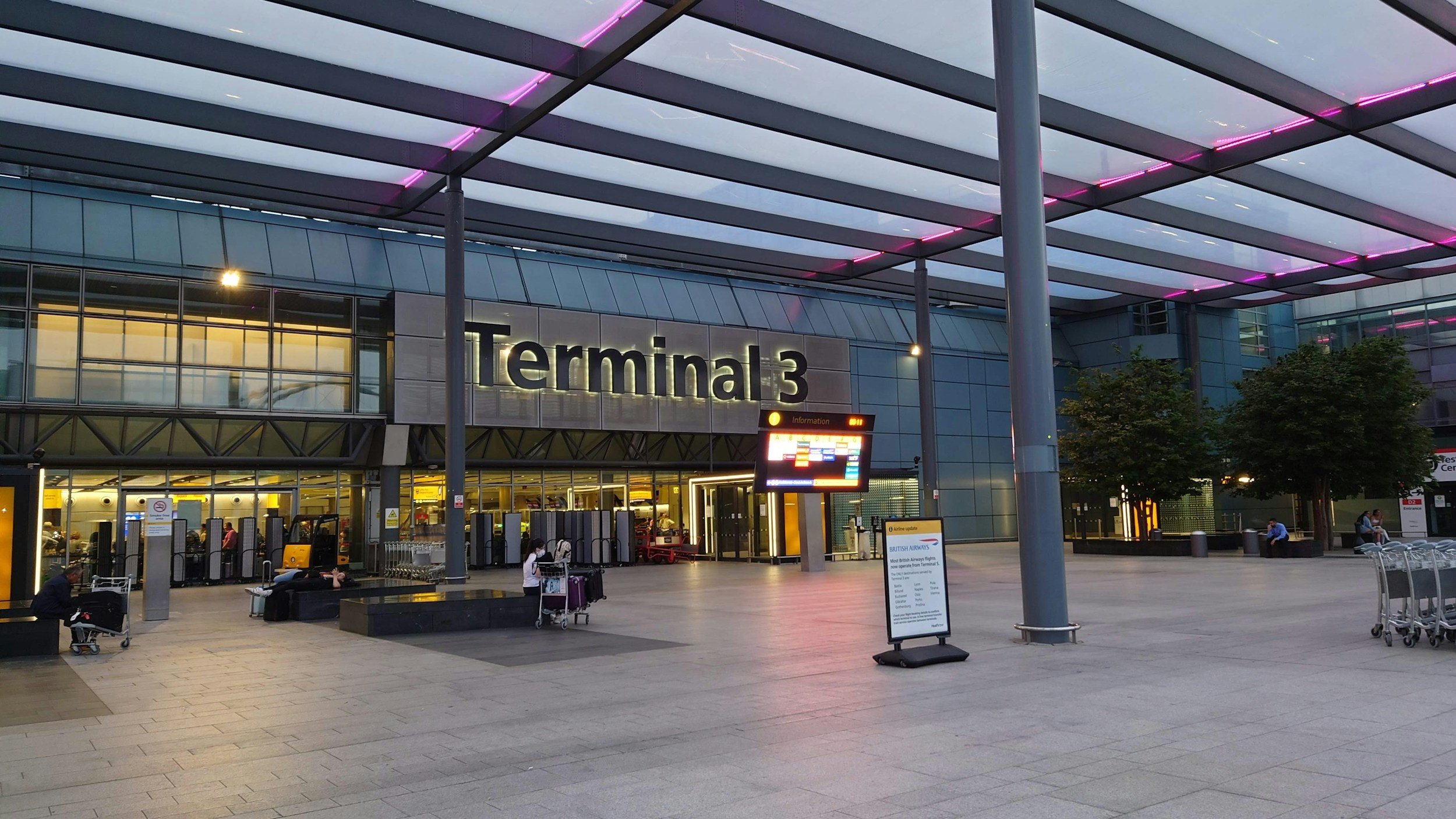 Exterior view of Terminal 3 at an airport during dusk, with glass walls, seating area, and a sign for British Airways.