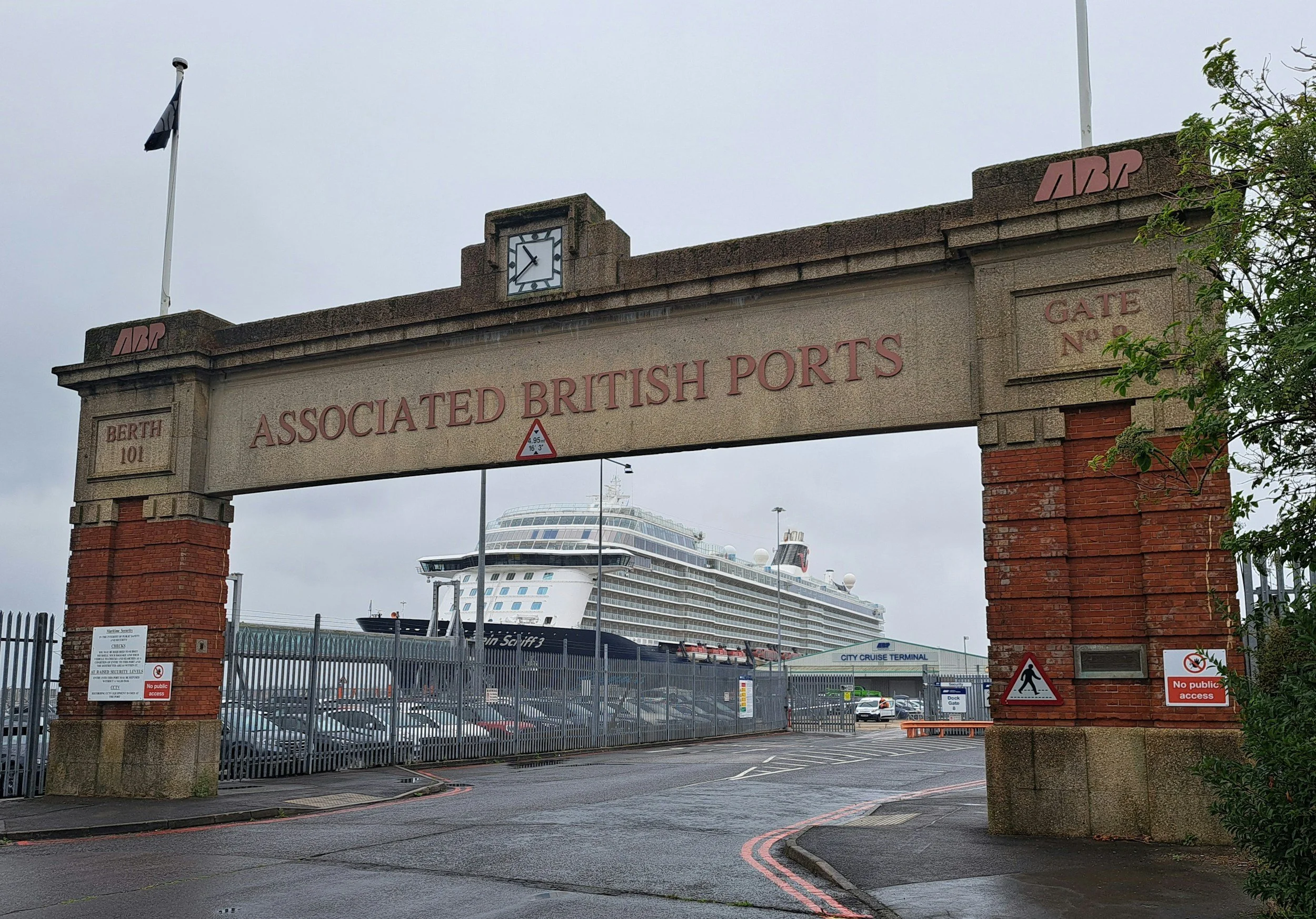 Gate entrance with brick pillars and a metal archway reading "Associated British Ports" leading to a parking lot with a cruise ship docked in the background.