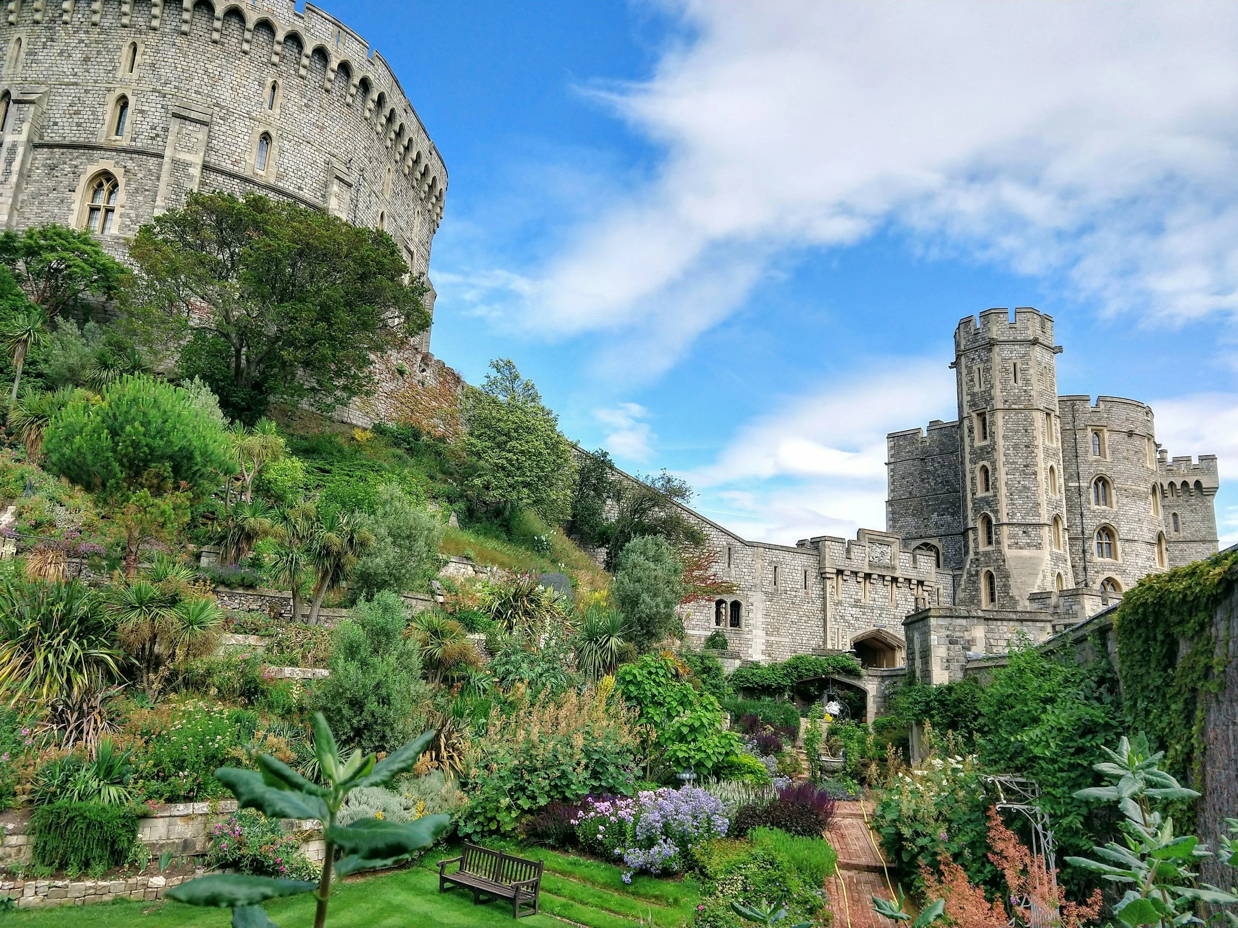 A castle with a round tower on the right and a larger rounded section on the left, surrounded by lush greenery and a colorful flower garden, under a partly cloudy blue sky.