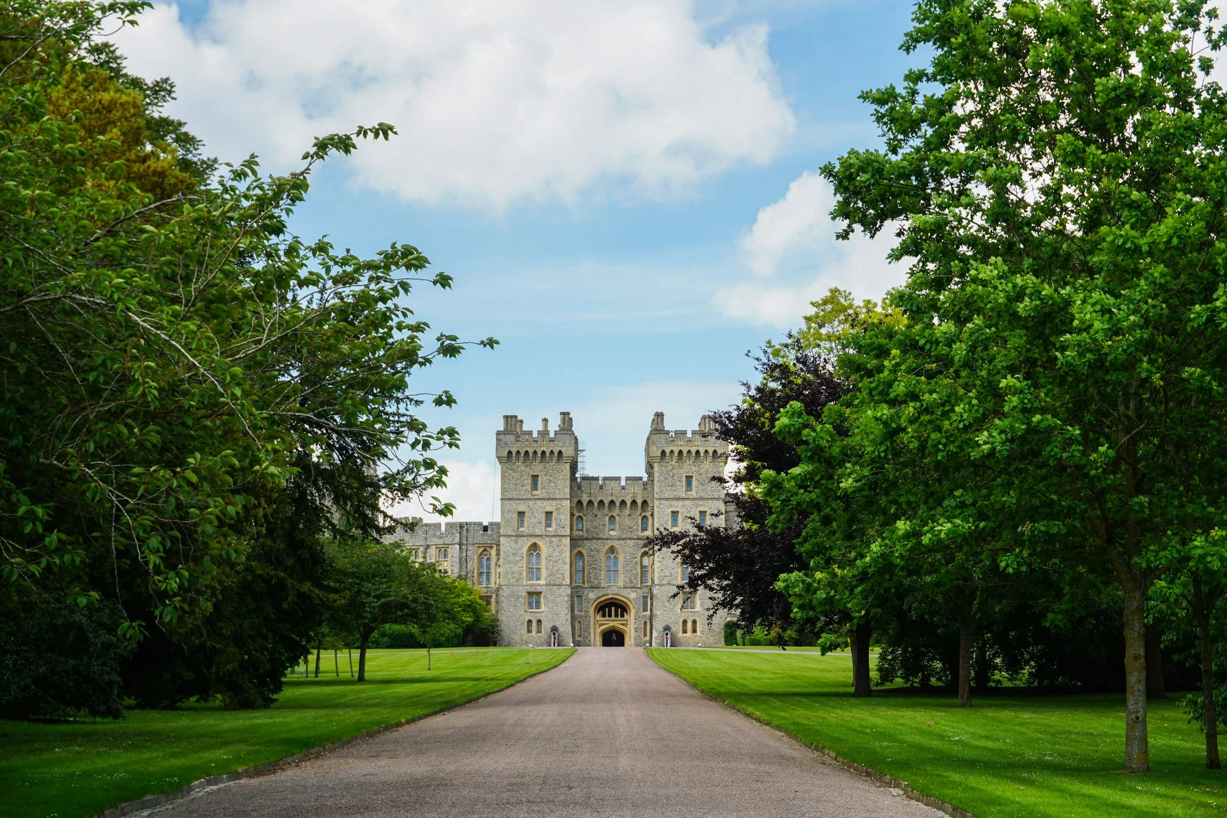 A castle surrounded by lush green trees and a manicured lawn, with a gravel driveway leading up to the entrance, under a partly cloudy sky.