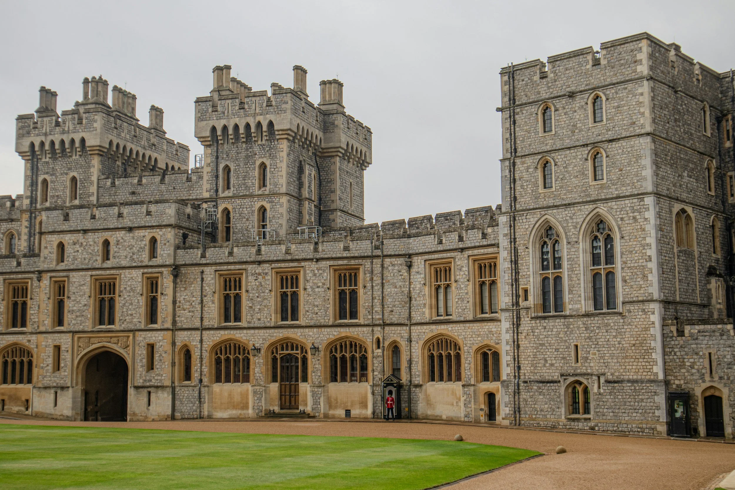 Front view of a medieval castle with stone walls and towers, a person in uniform standing near the entrance, and a green lawn in the foreground.